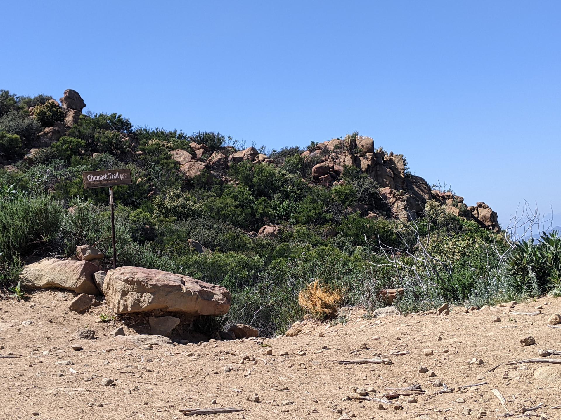 A trail sign next to large rocks on a sunny, rocky hillside.