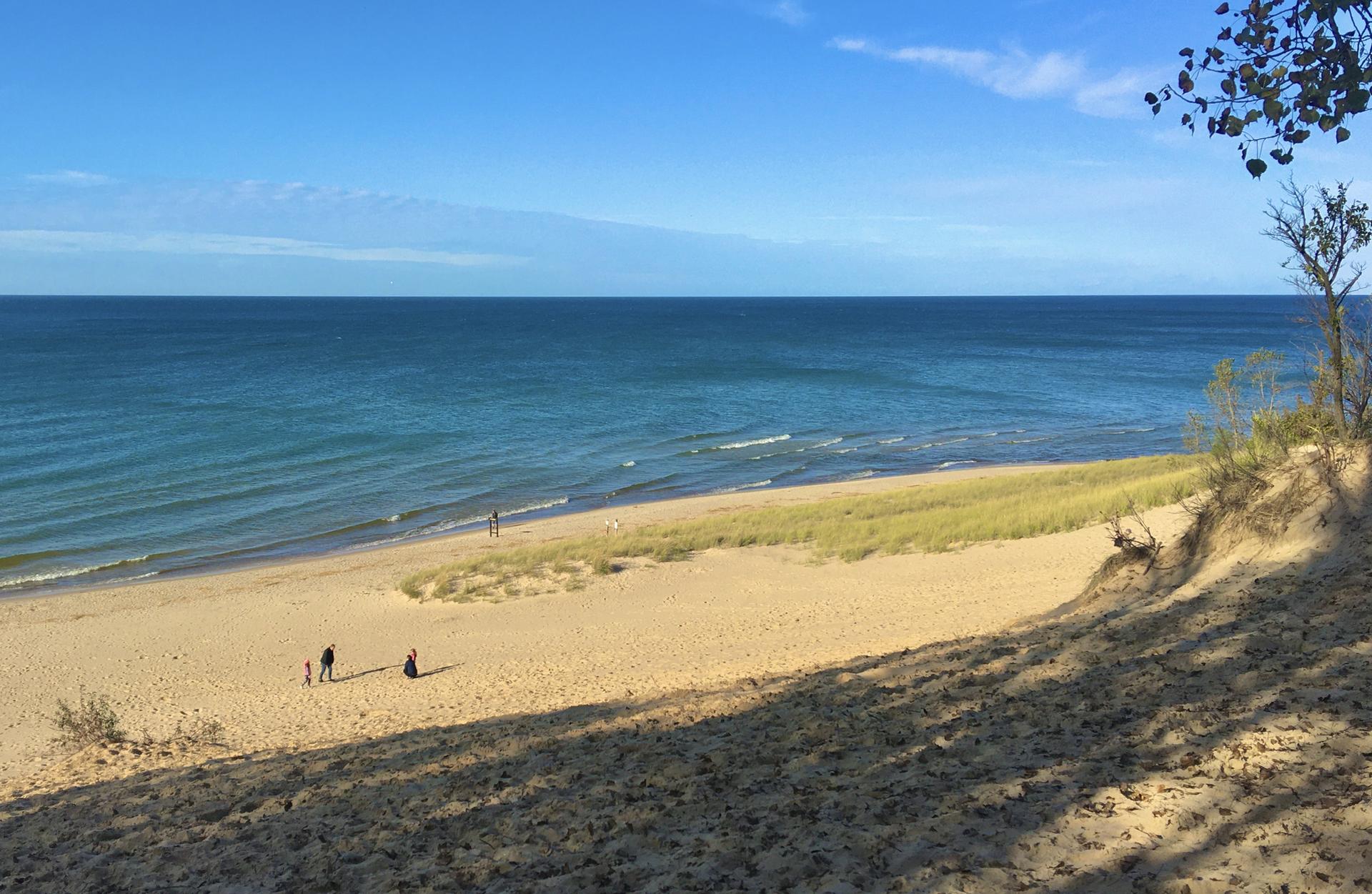 Sunny beach with blue ocean, sandy dunes, and distant people walking.