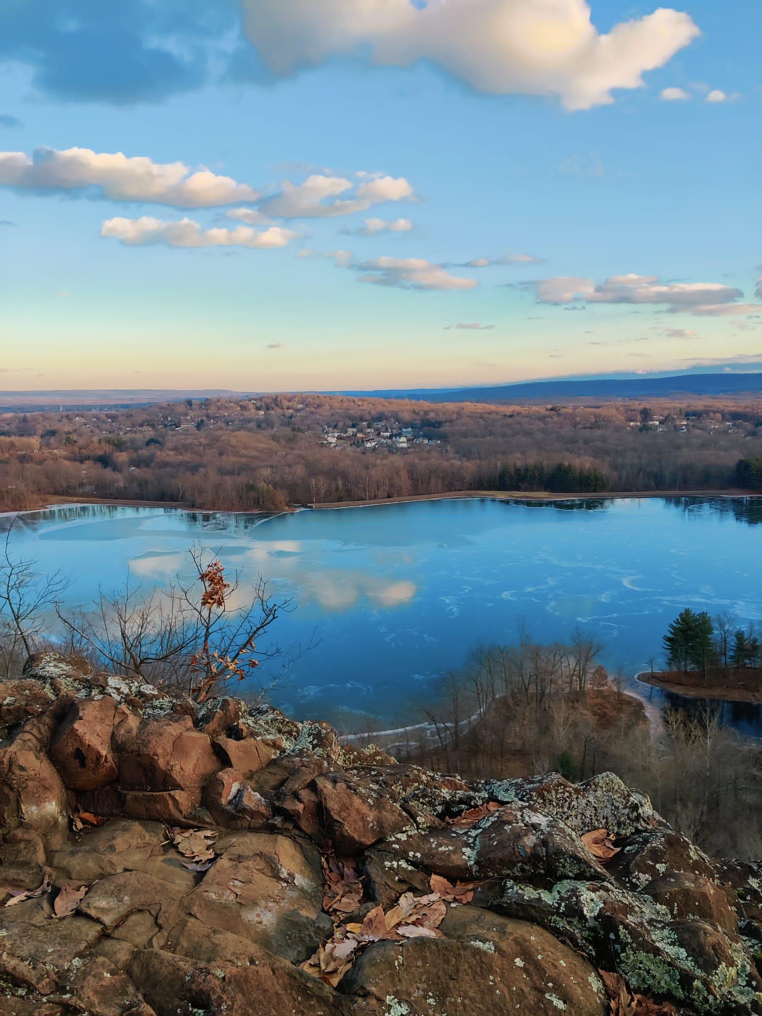Overlook of a partially frozen lake surrounded by bare trees under a cloudy sky.