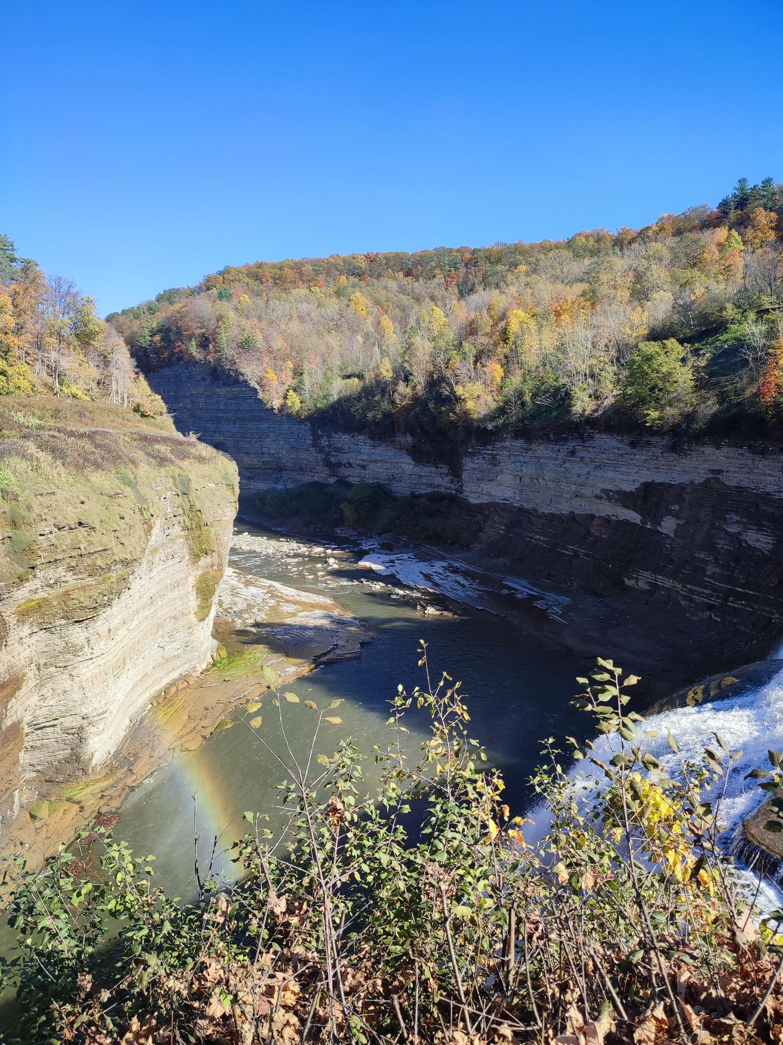 Sunny autumn gorge with a river, colorful trees, and a small rainbow.