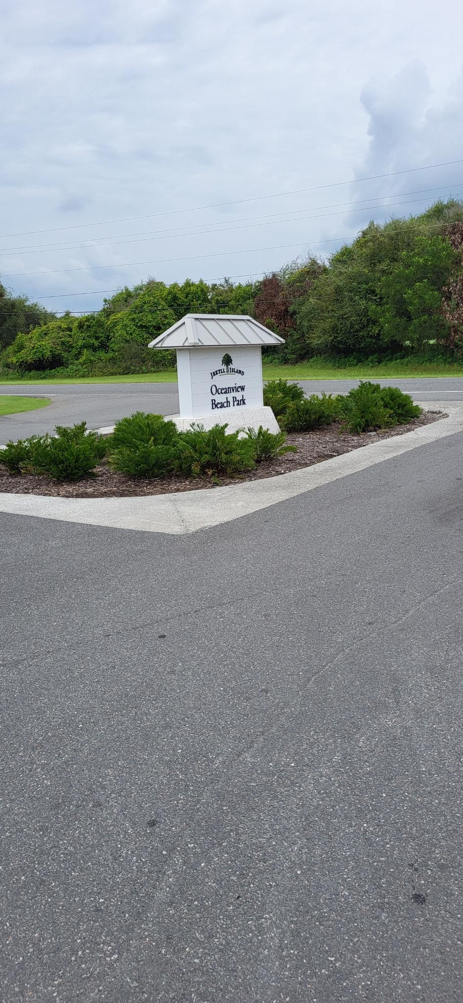 A white park sign surrounded by bushes at a paved road intersection.