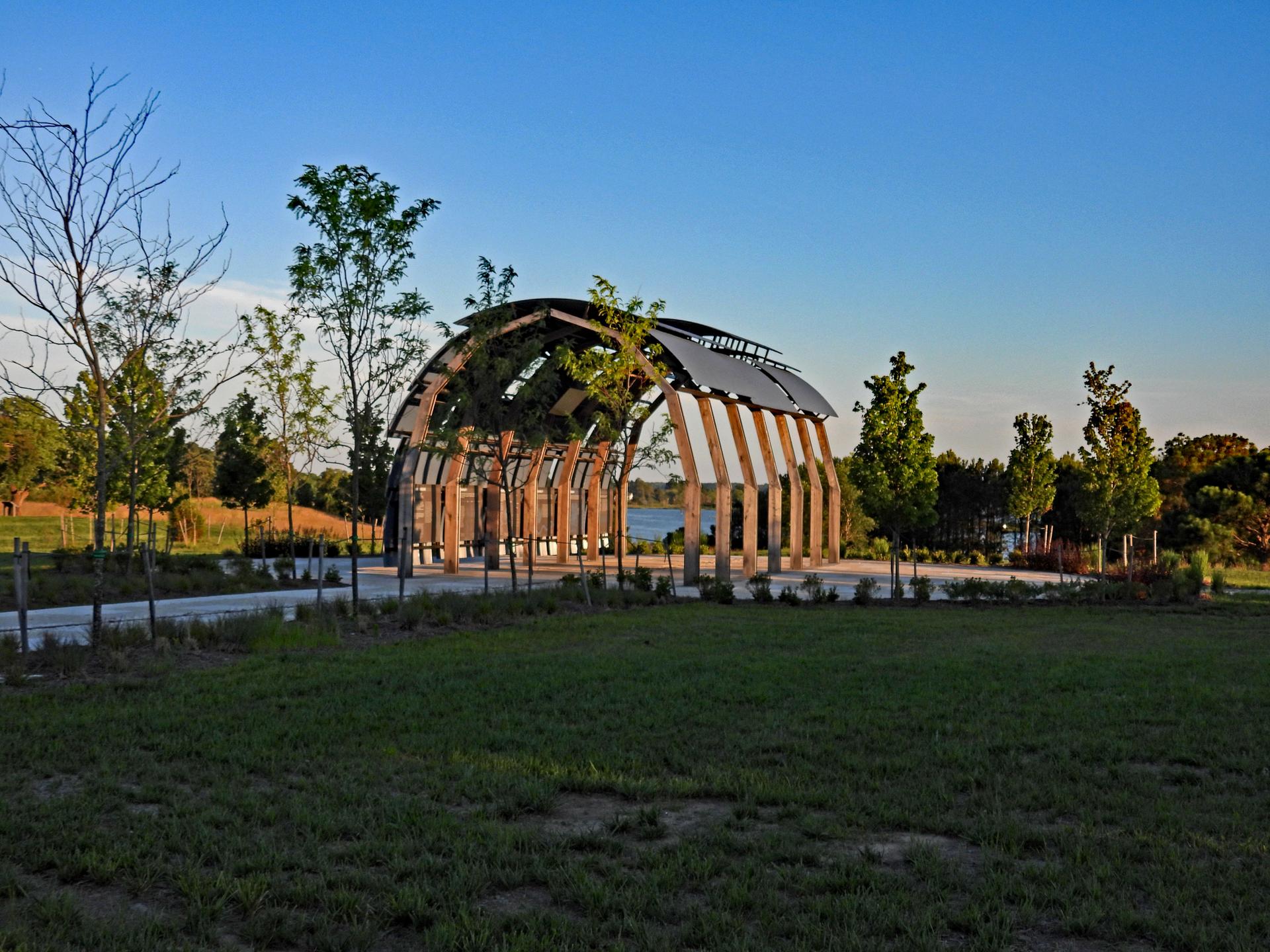 Modern arched wooden pavilion by a lake, surrounded by trees under a clear sky.