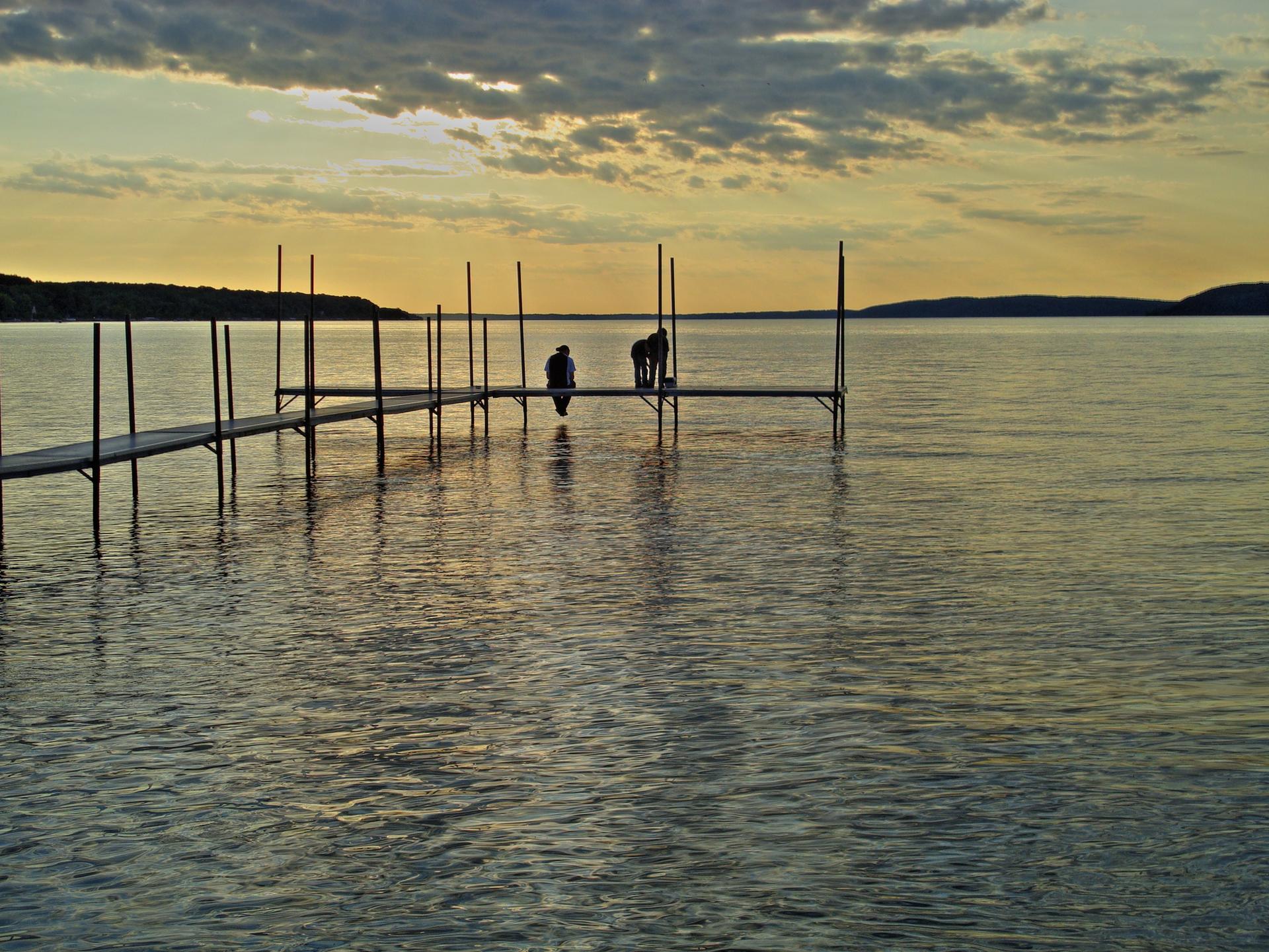 Two people on a long dock over a calm lake at sunset.