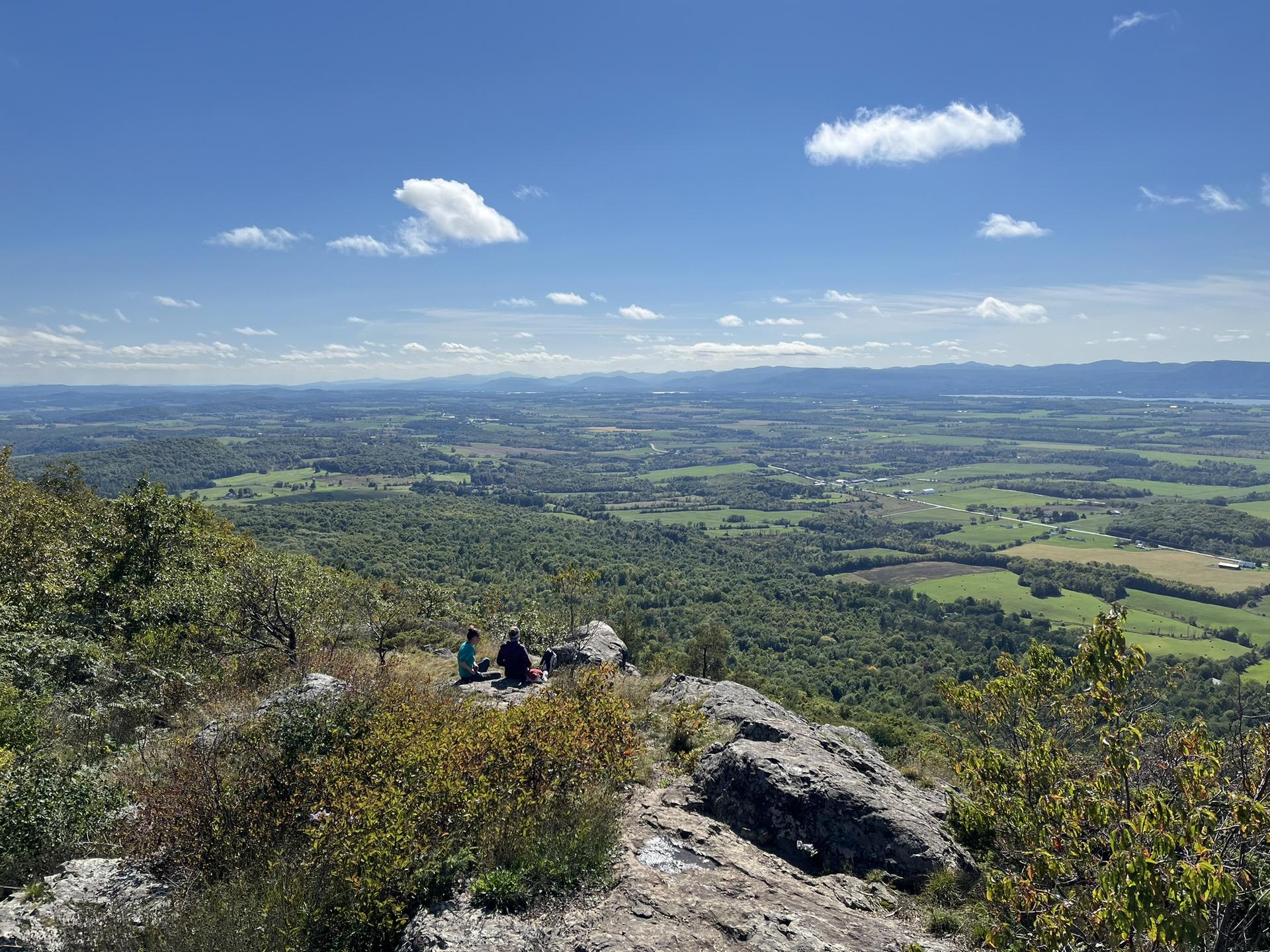 People on a rocky peak overlook a vast green valley under a sunny sky.