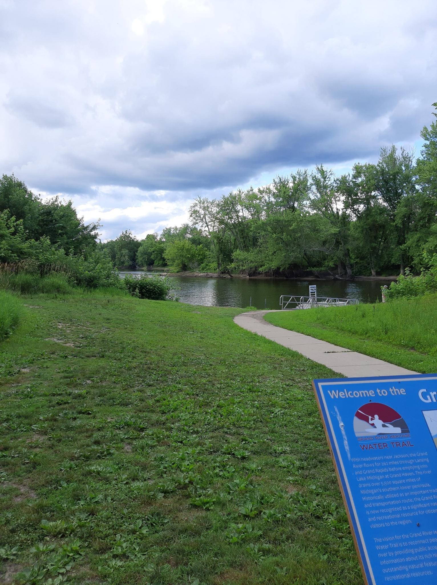 A paved path leads to a calm river surrounded by dense green trees.