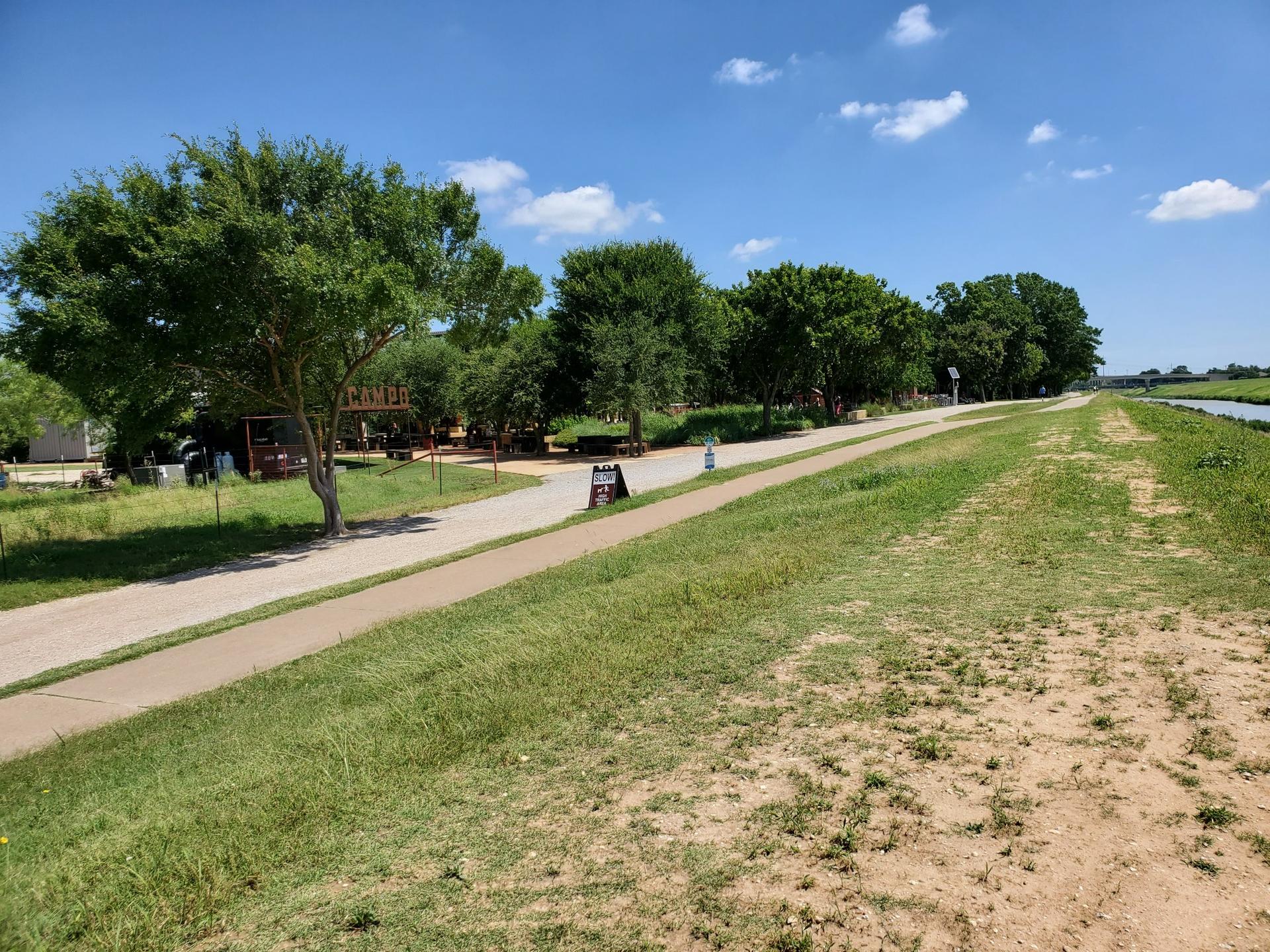 Paved path next to a grassy field and trees on a sunny day.