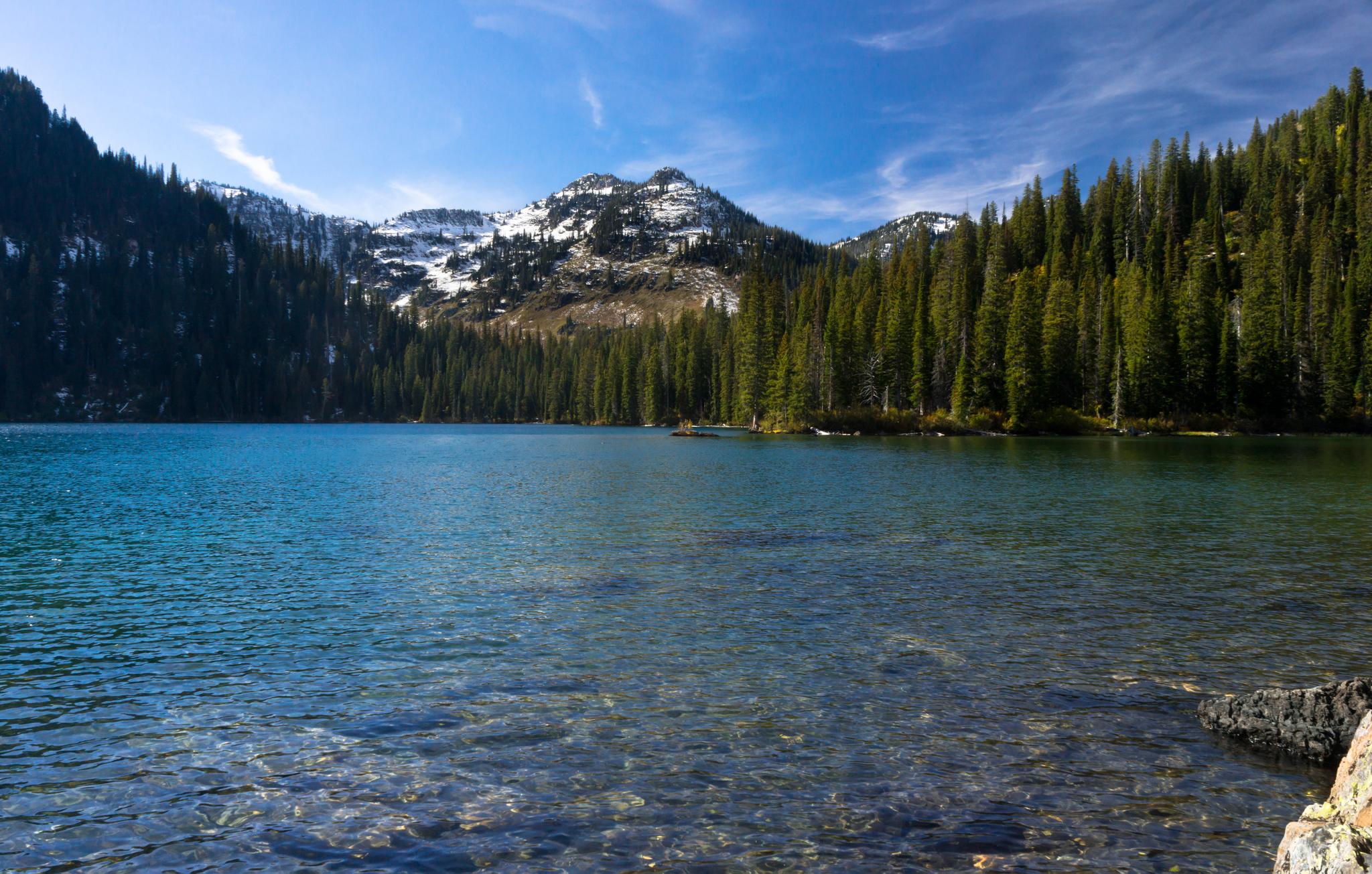 Clear mountain lake surrounded by evergreen forests and snow-capped peaks under a bright sky.