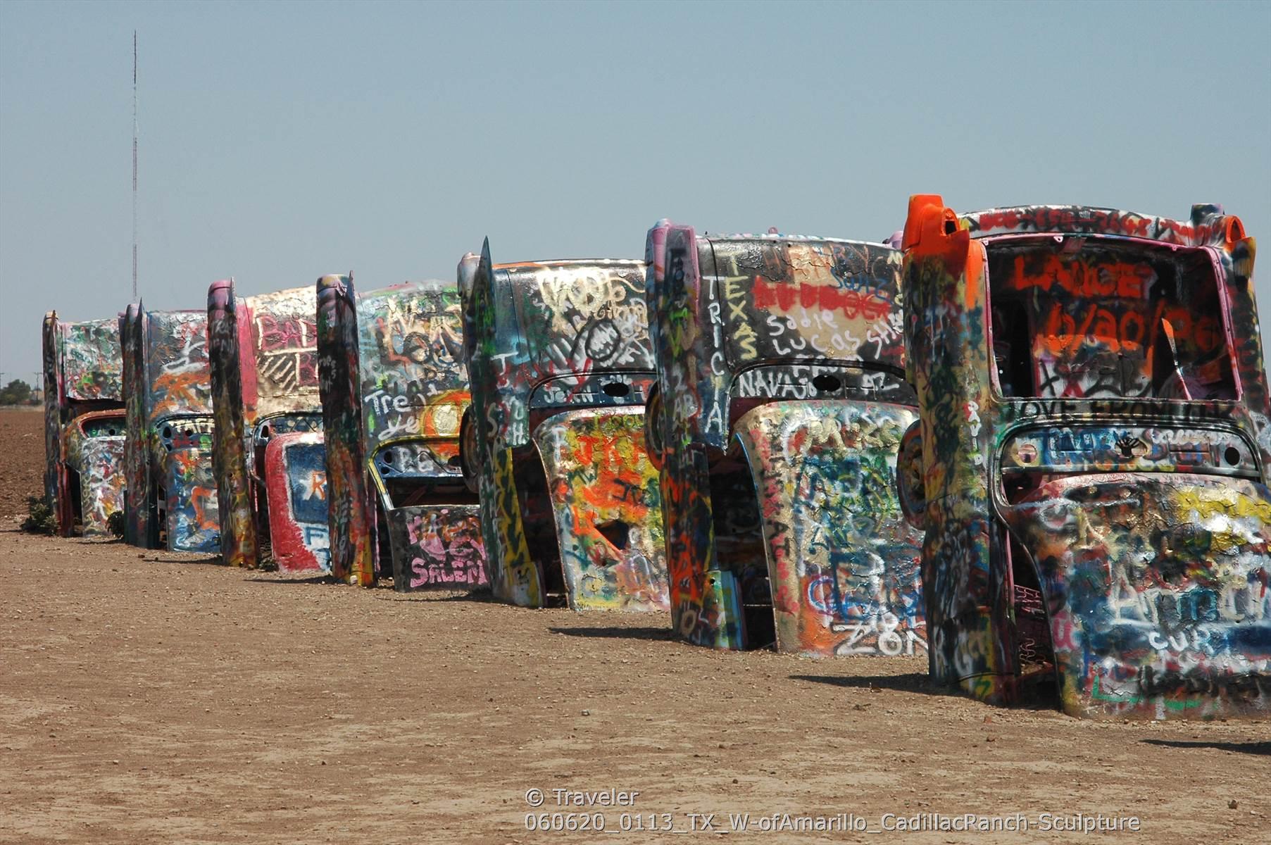 Row of graffiti-covered cars buried nose-down in dry, flat ground under bright sky.