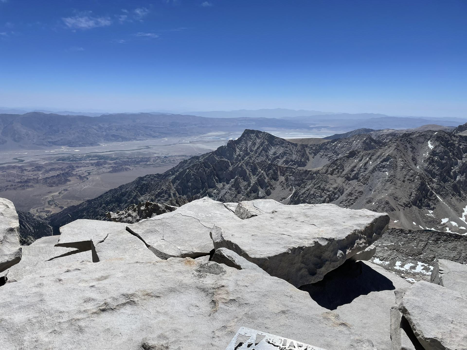Rocky mountain peak with vast valley and distant mountains under a clear sky.
