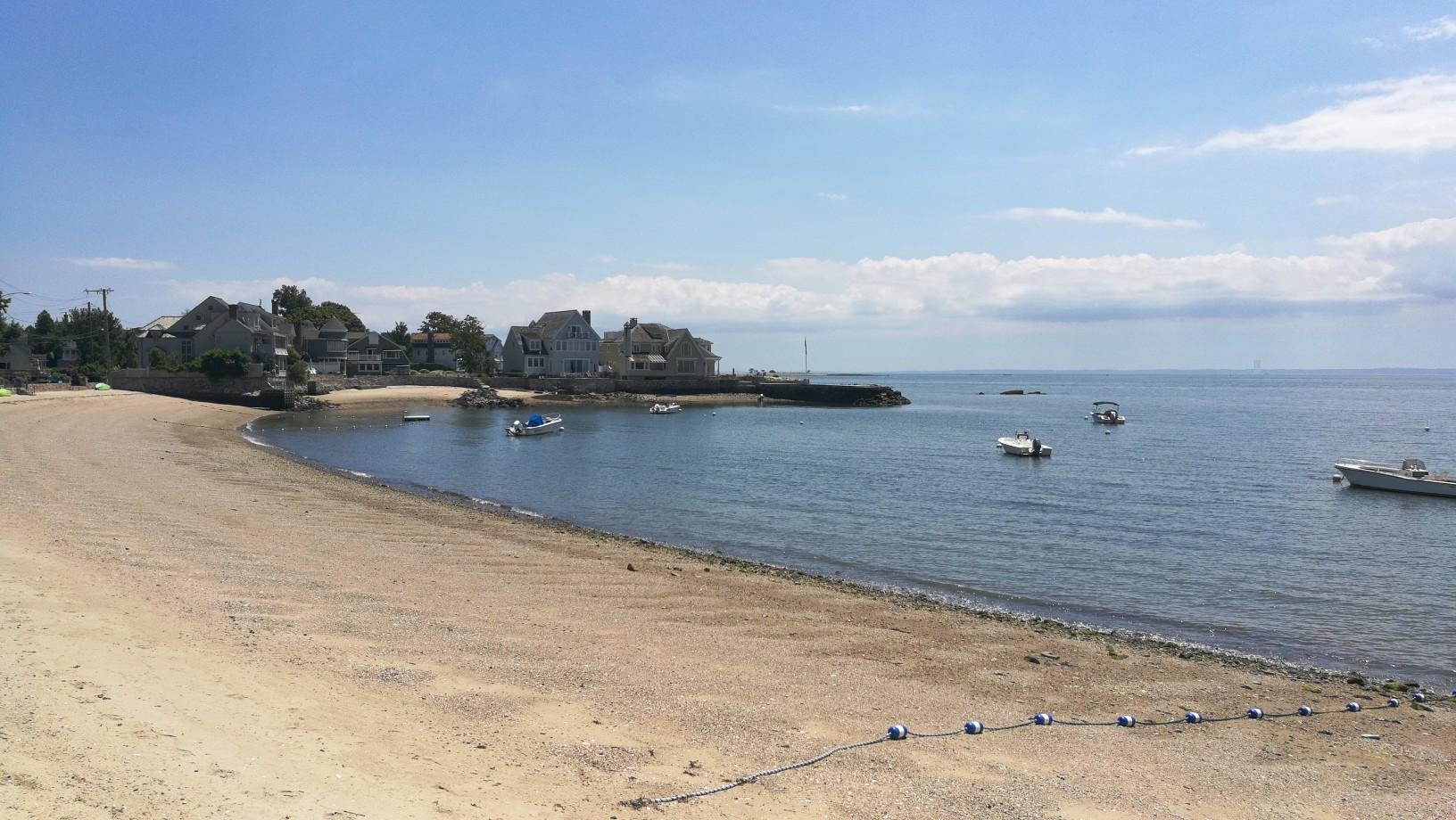 Sunny beach with boats on blue water and houses along the shore.