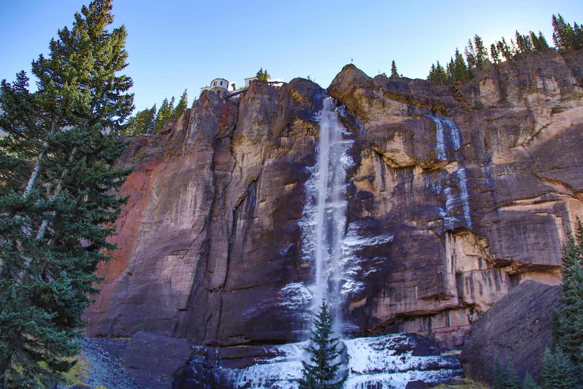 Powerful waterfall cascades down a massive rocky cliff, surrounded by pine trees.