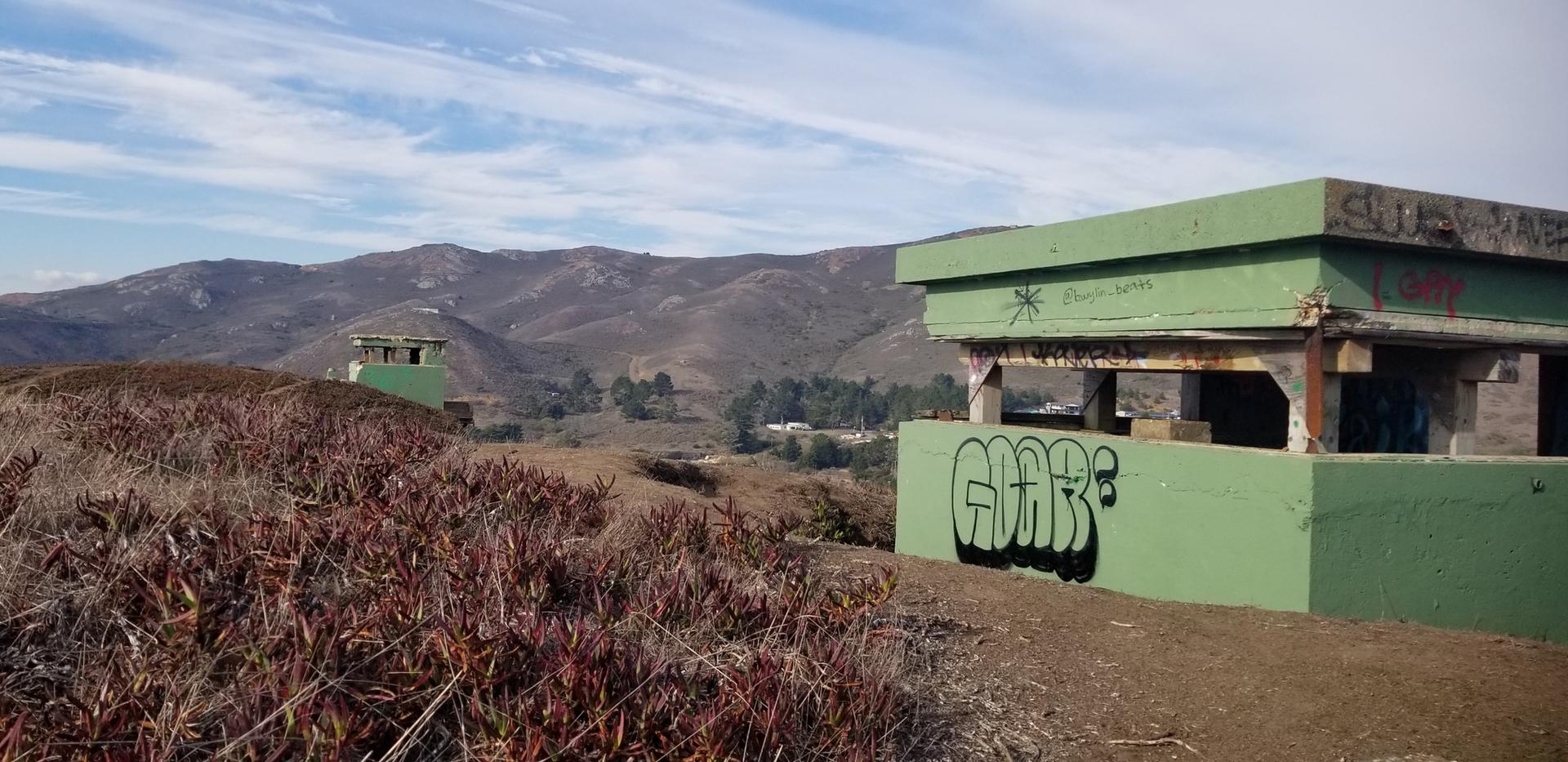 Green concrete structures with graffiti on a hillside, mountains in background, sunny.