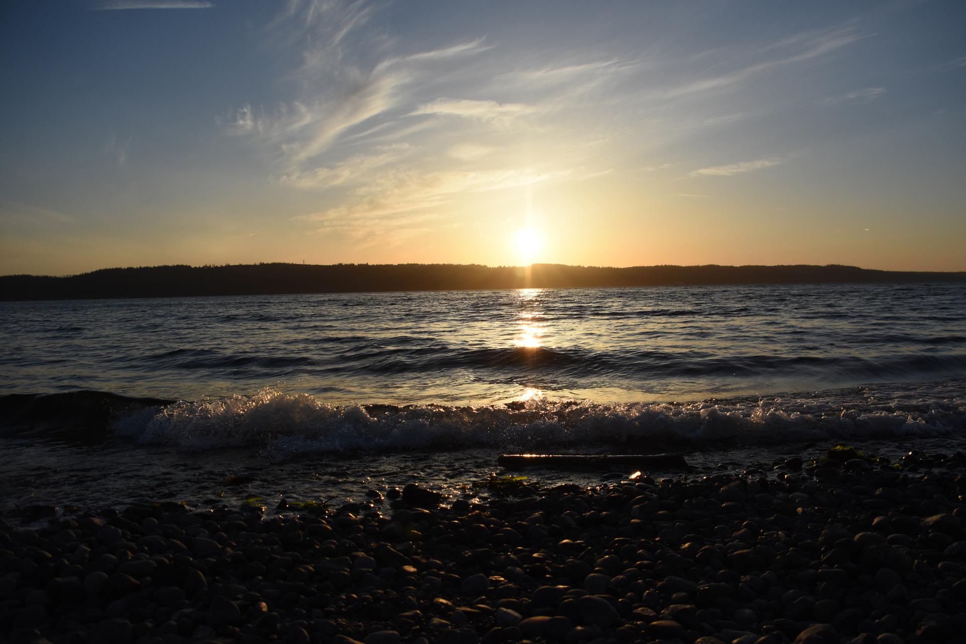 Sunset over a calm body of water with waves breaking on a rocky shore.