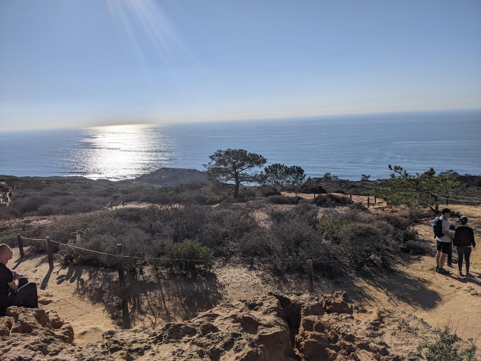 People on a sunny coastal trail with ocean views and sparkling water.