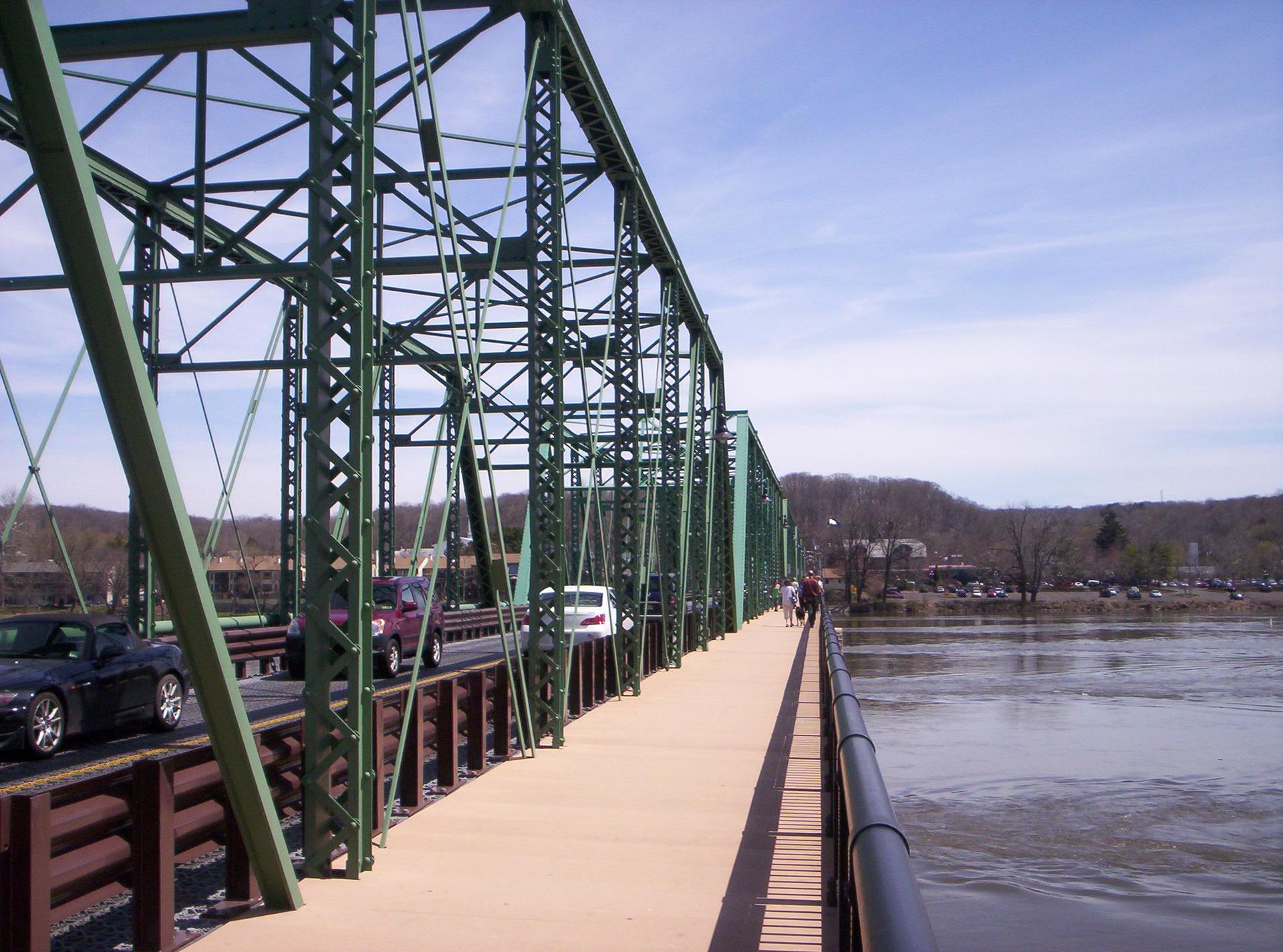 Green metal truss bridge with cars and pedestrians, spanning a river on a sunny day.