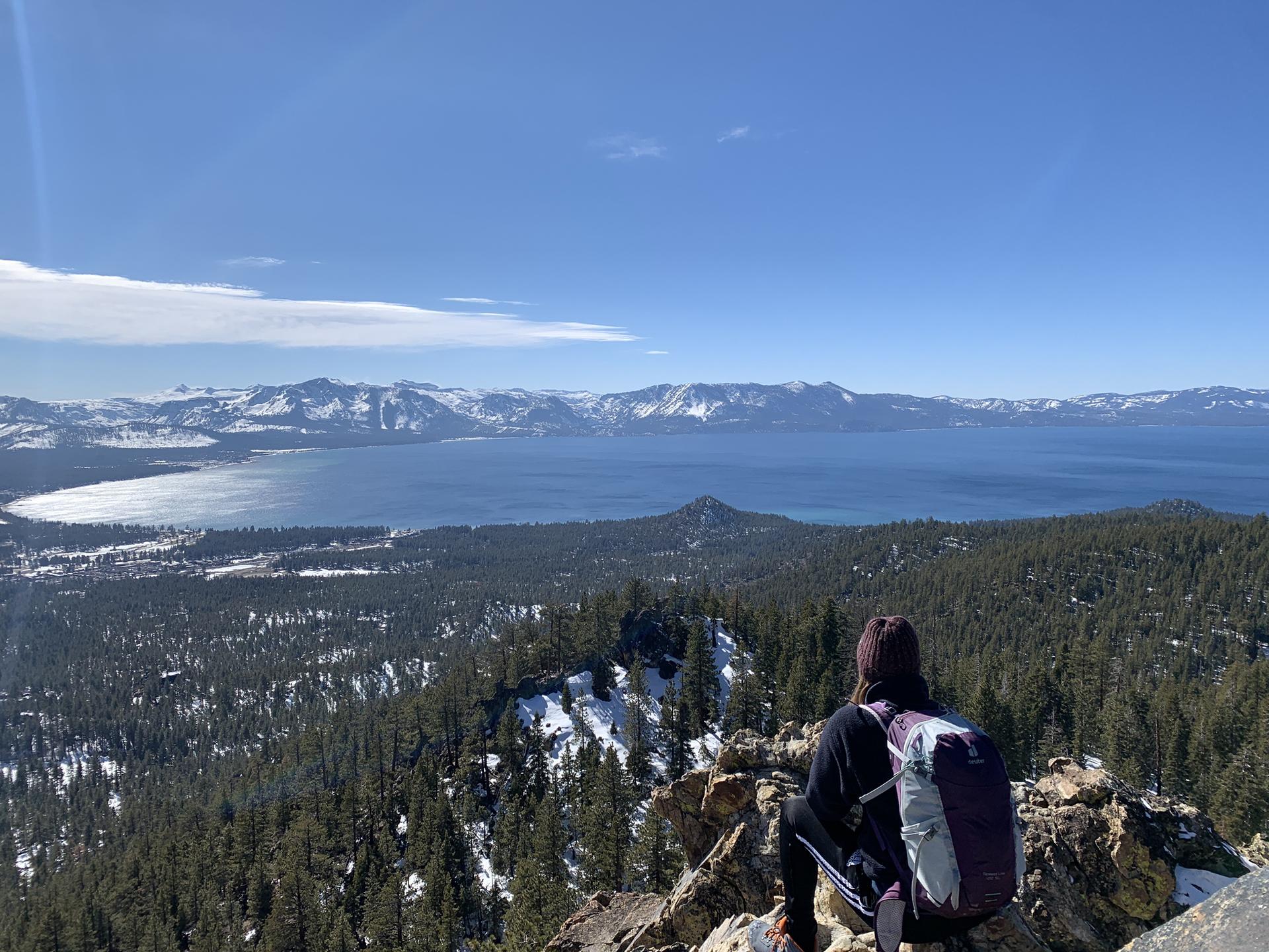 Person with backpack sits on rocks, overlooking a vast lake and snowy mountains.