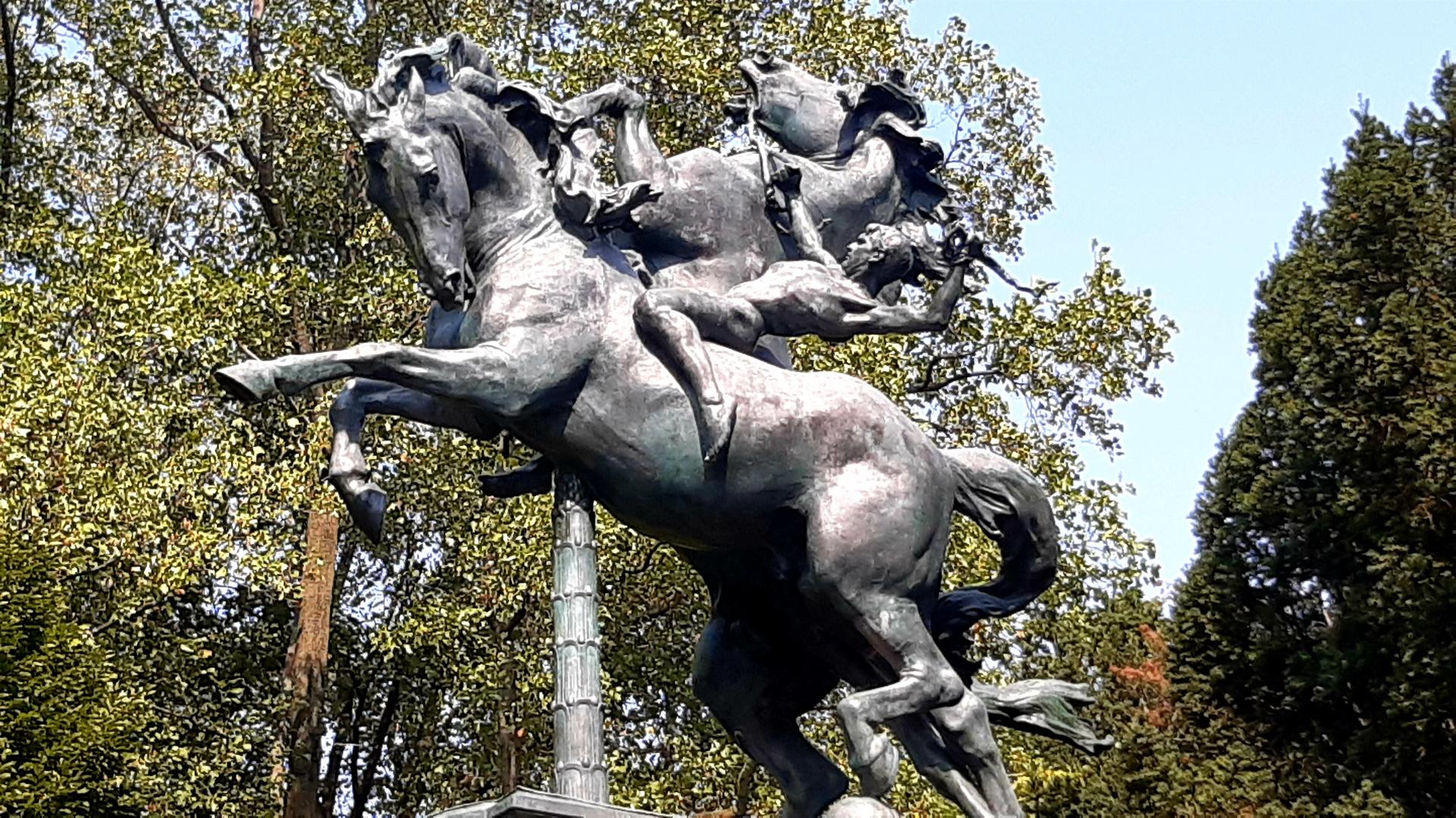 Bronze statue of a rearing horse with a figure, surrounded by green trees.