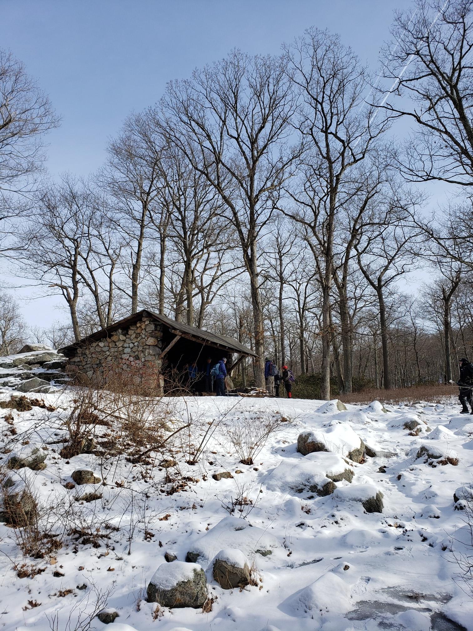 Stone shelter in a snowy forest with tall bare trees and people.