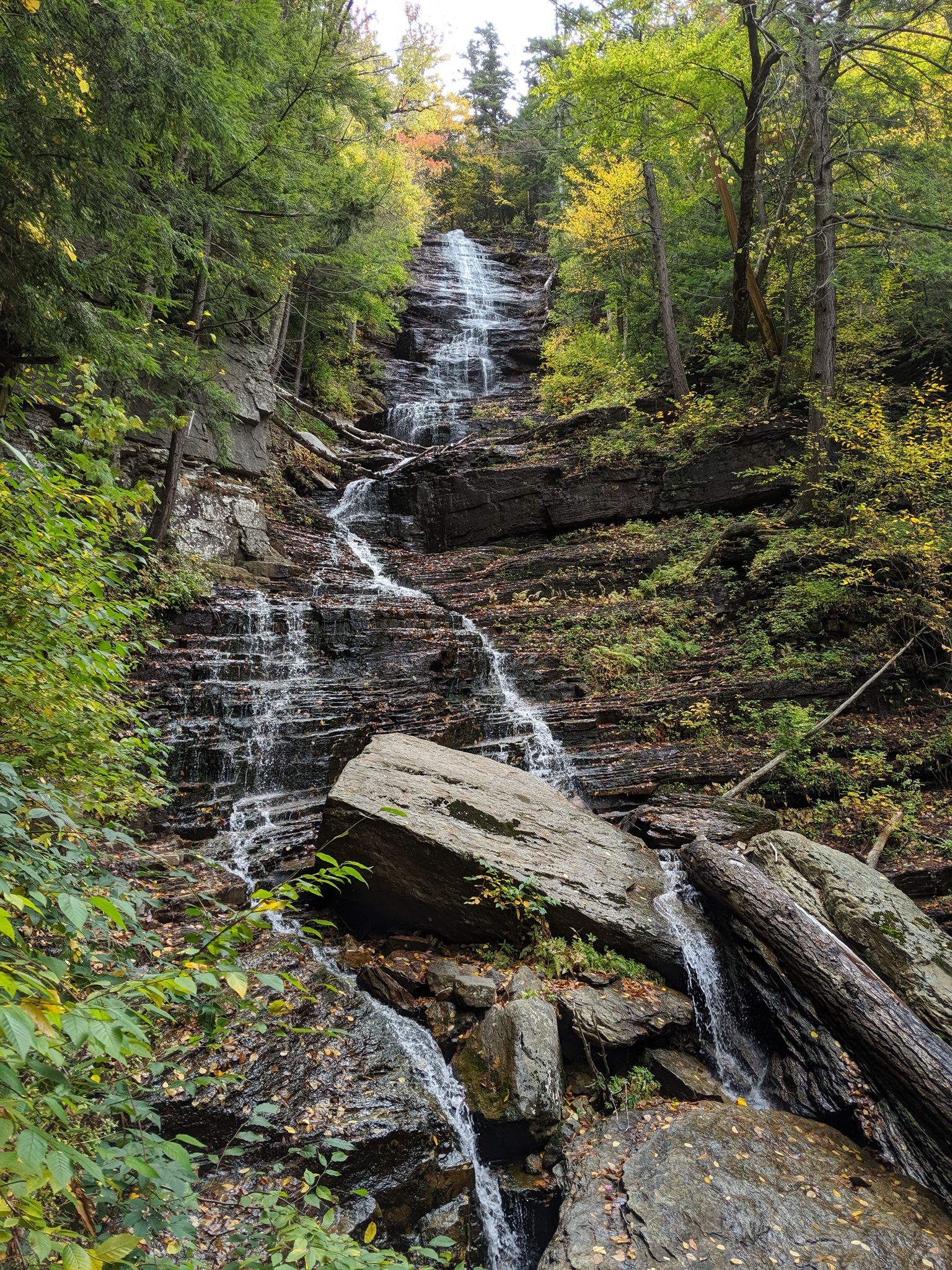 A tall, multi-tiered waterfall cascades down rocky cliffs surrounded by lush forest. - Lye Brook Falls Trail