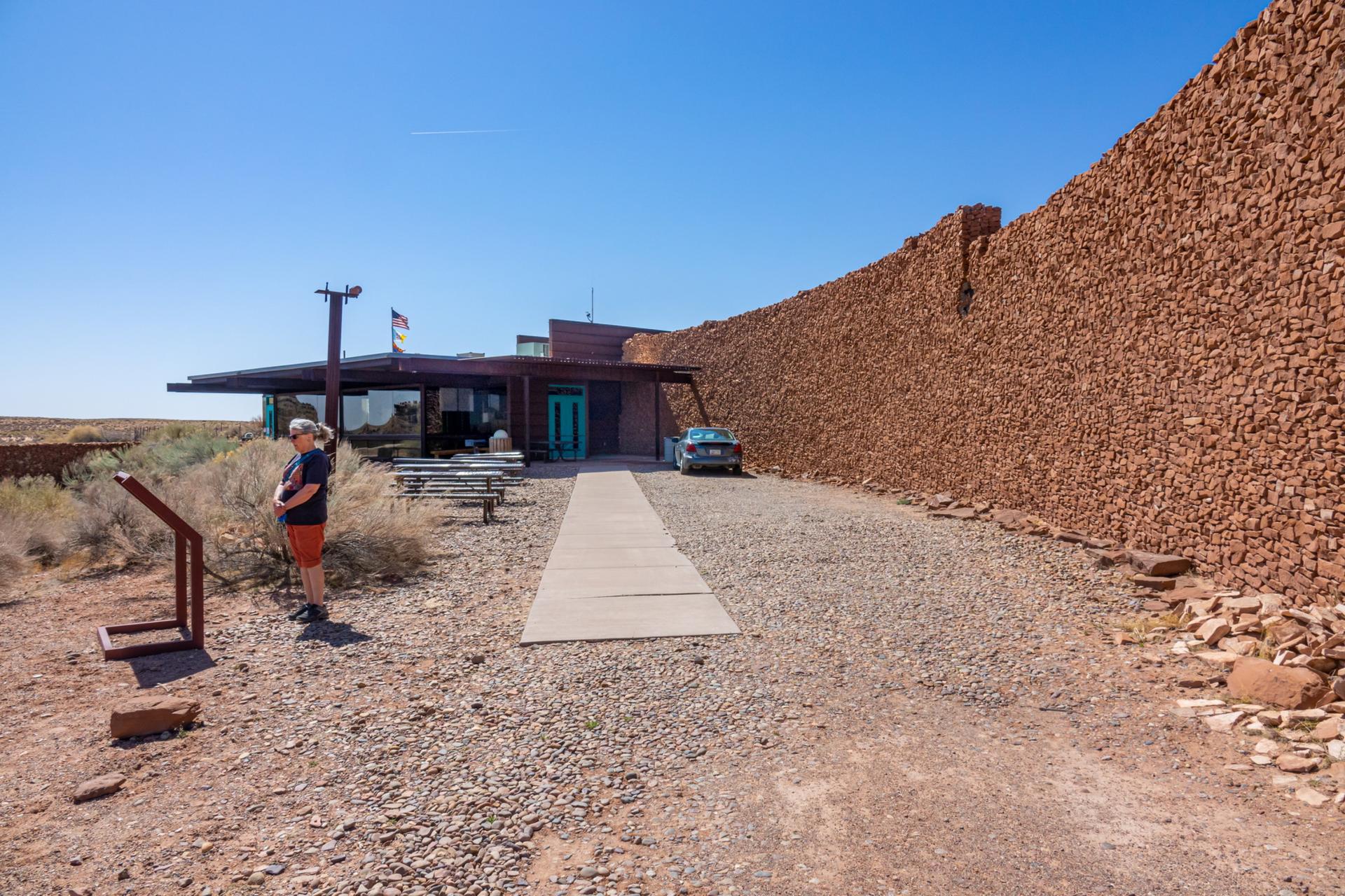 Person stands on gravel near a building and large stone wall under clear sky.