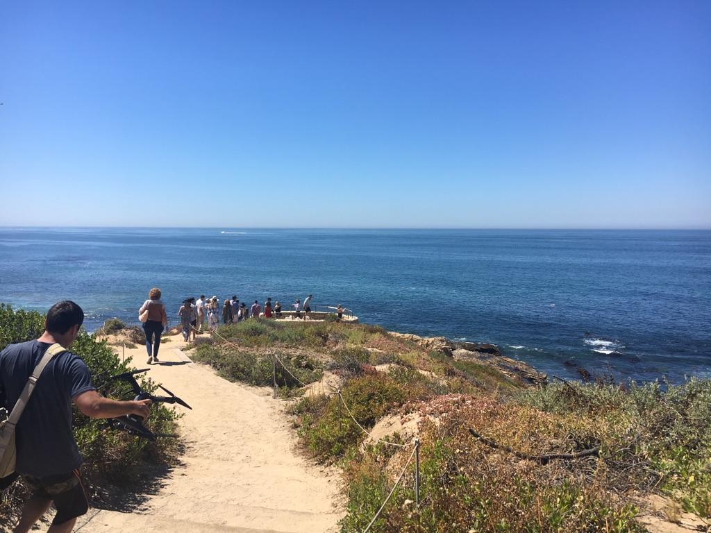 People walk on a sunny dirt path overlooking the vast blue ocean.
