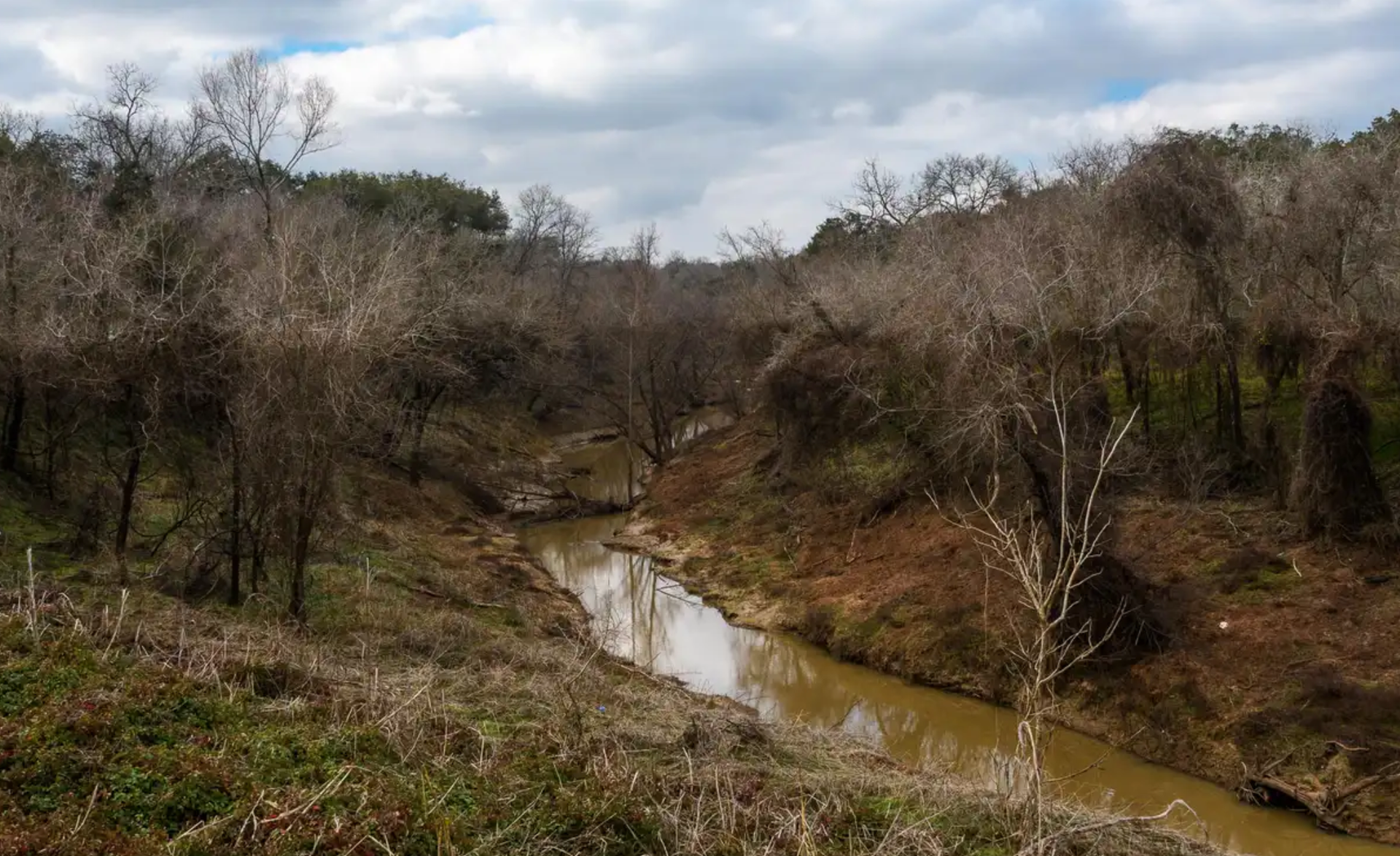 A muddy river winds through a valley with bare trees and brown foliage under a cloudy sky.