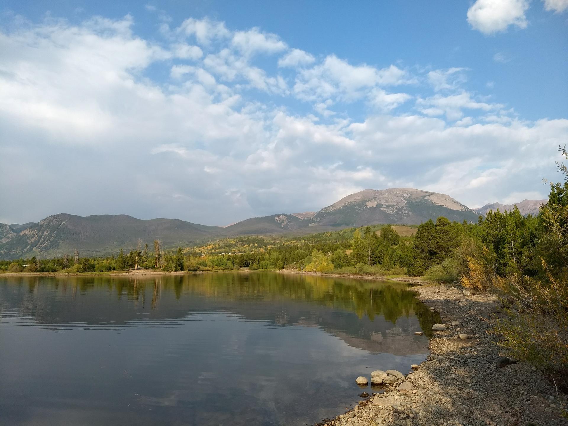A clear lake reflects a partly cloudy sky with forested mountains.