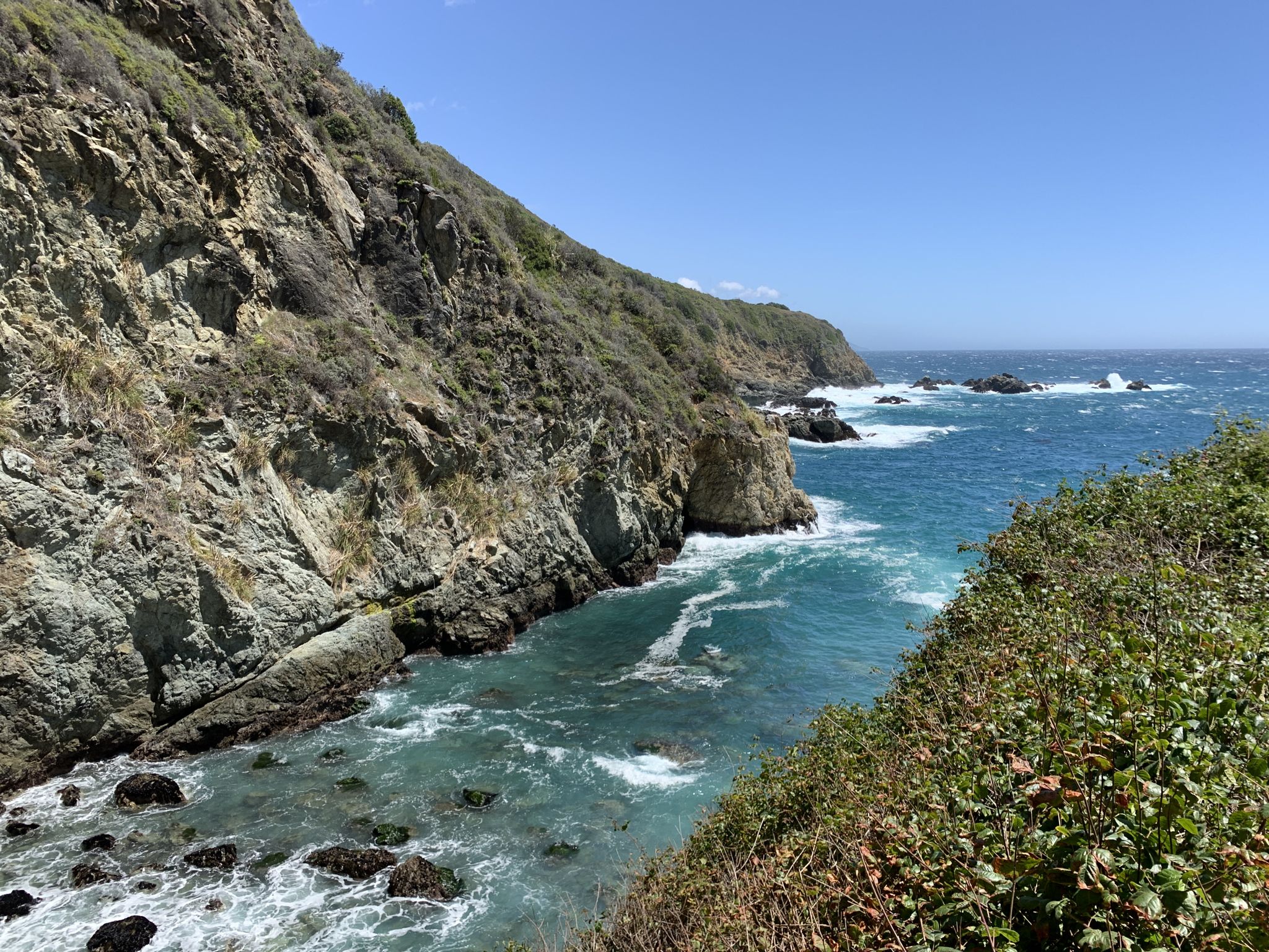 Steep, green cliffs meet a blue ocean with white waves under clear sky. - Partington Cove Trail