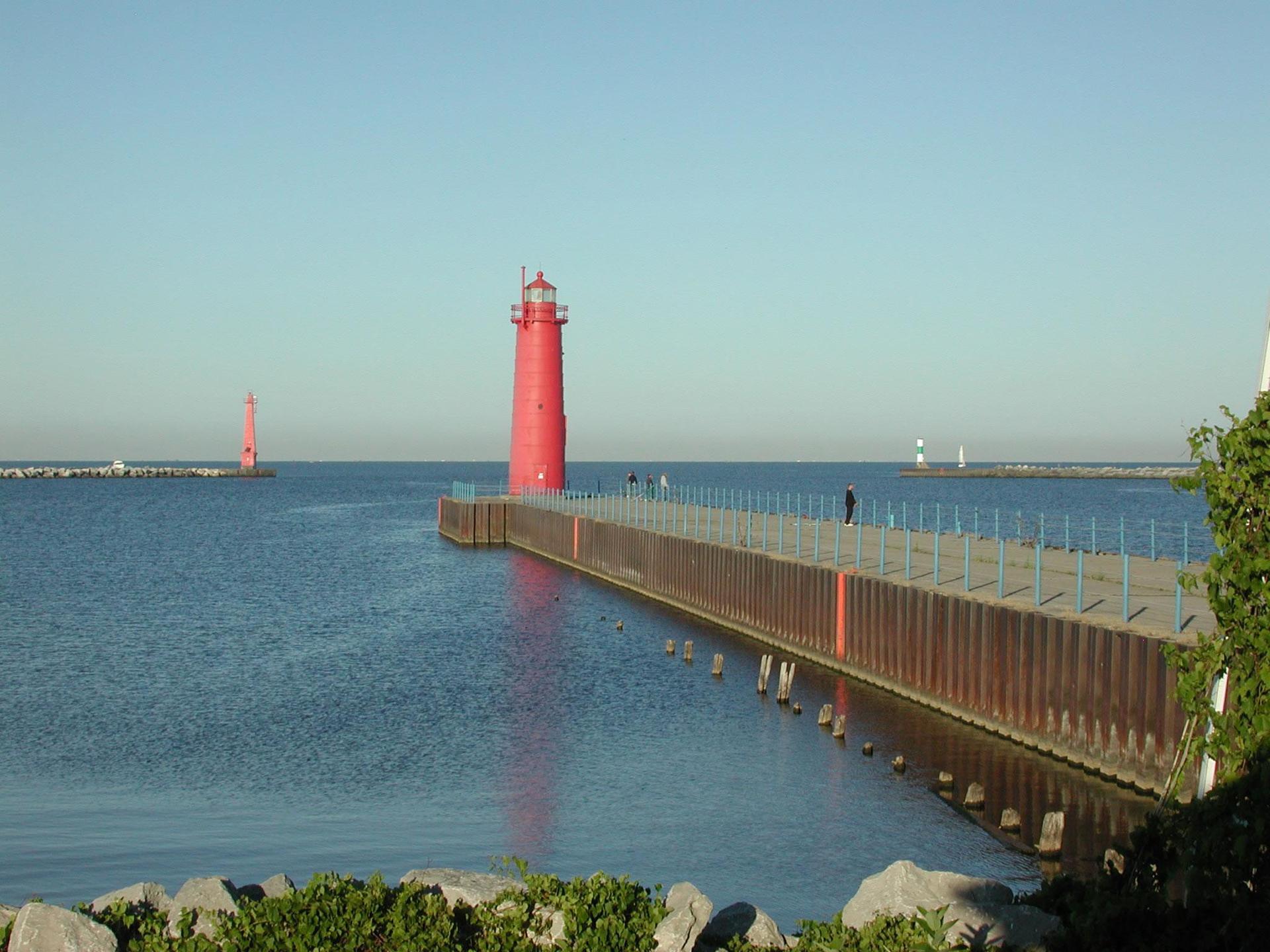 Red lighthouse and pier on calm blue water under a clear sky.