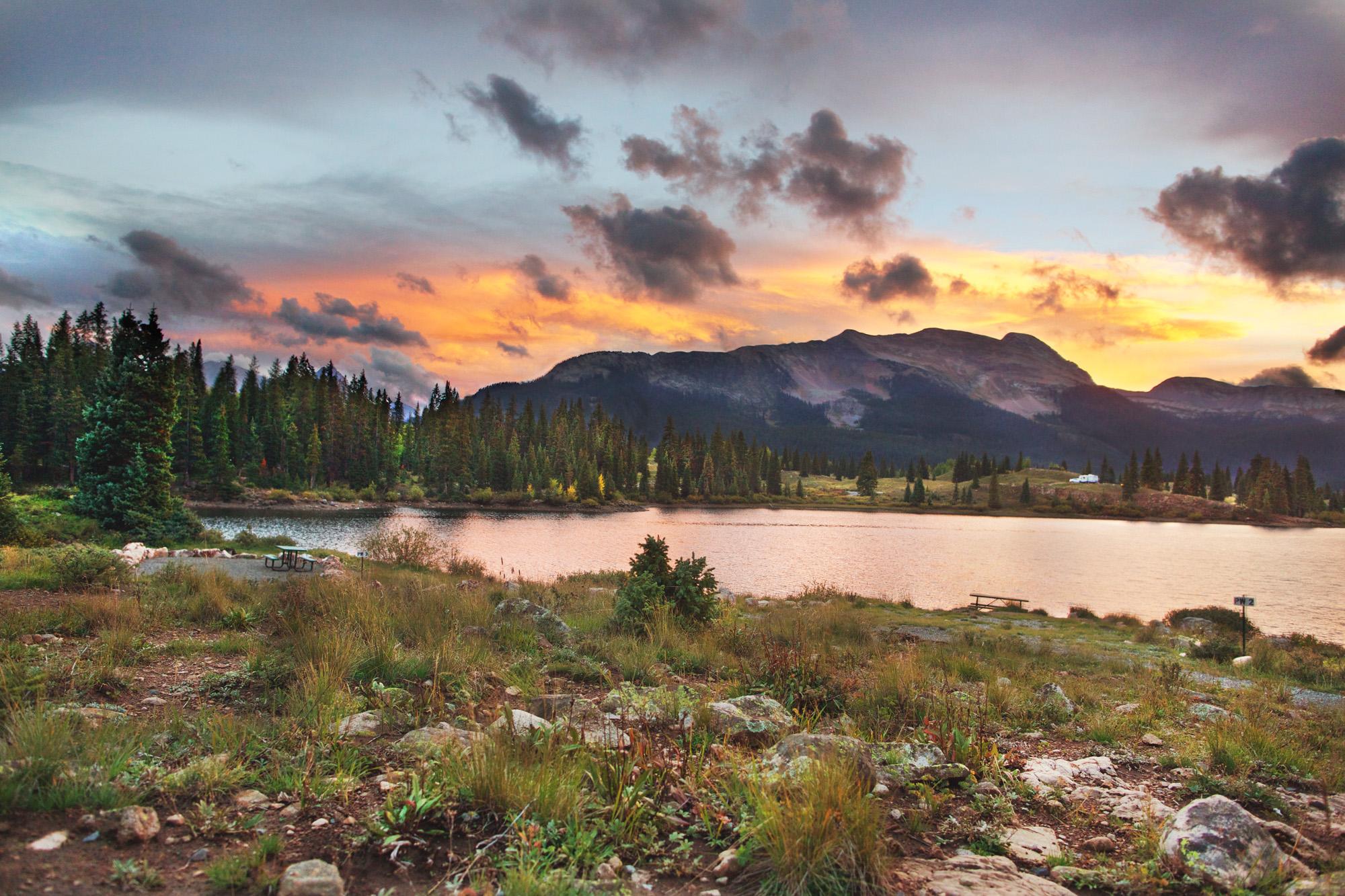 Lake at sunset with colorful clouds over mountains and dark forest.