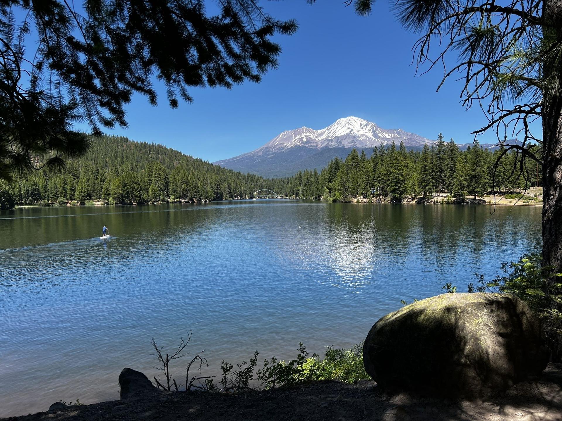 Serene lake reflecting a snow-capped mountain and forest on a sunny day.