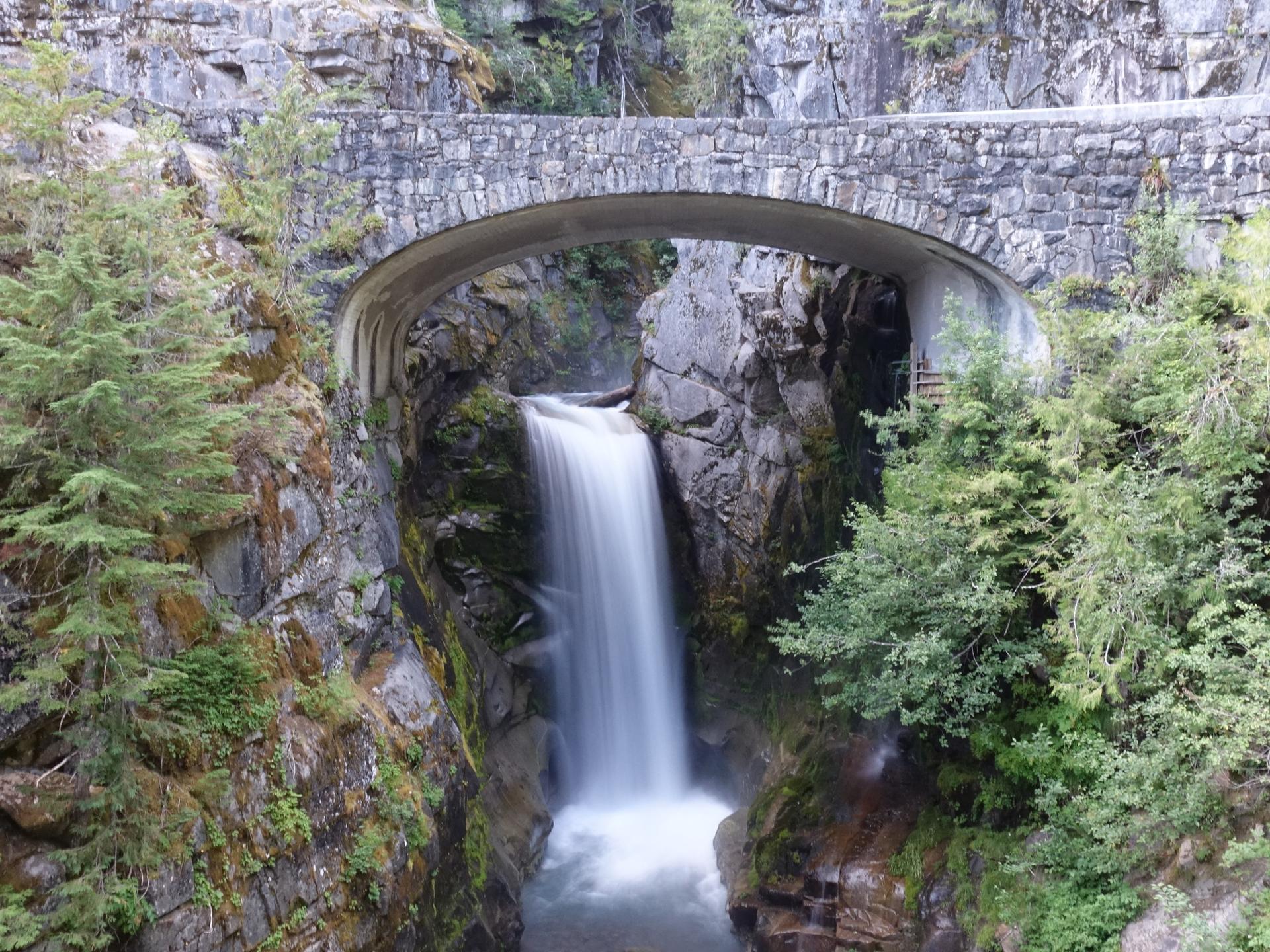 Waterfall flowing under a stone arch bridge in a rocky canyon.