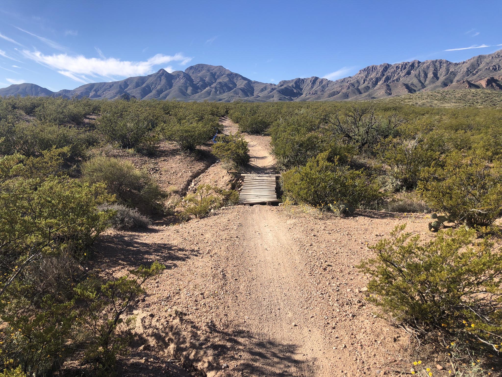 Desert path descends to a wooden bridge with mountains in the distance.