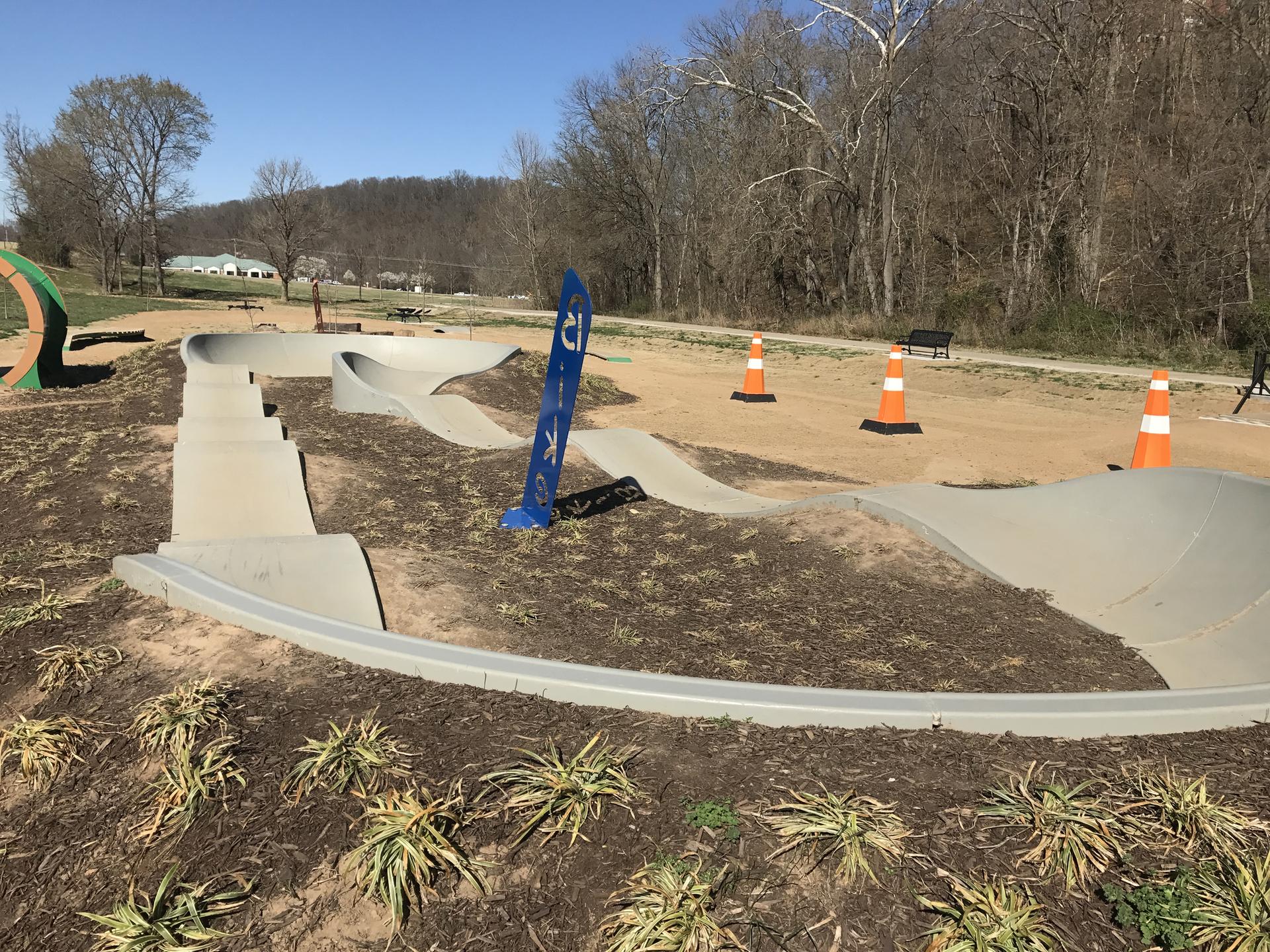 Concrete pump track under bright sun, surrounded by dirt and bare trees.