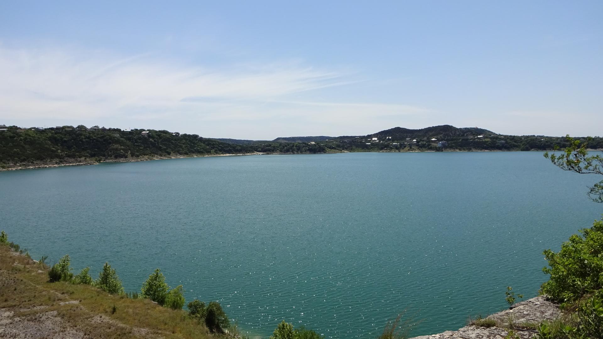 Expansive blue lake surrounded by green hills under a bright, clear sky.