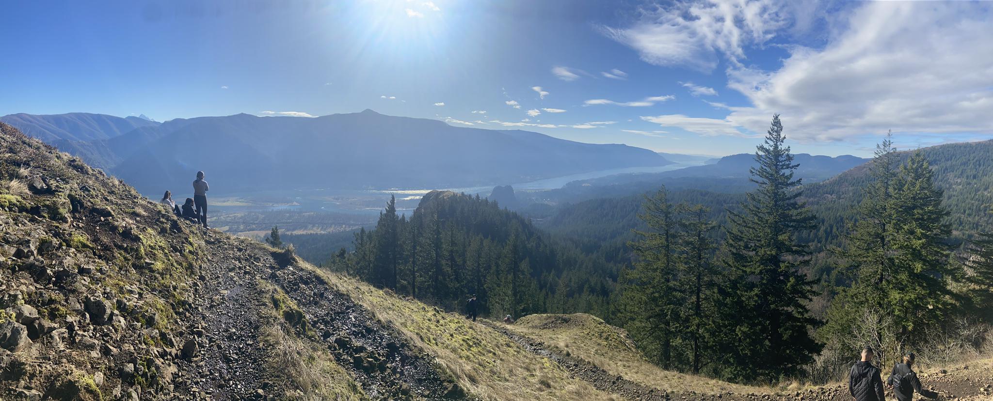 Vast valley, mountains, and forests under bright sun with people on trail.