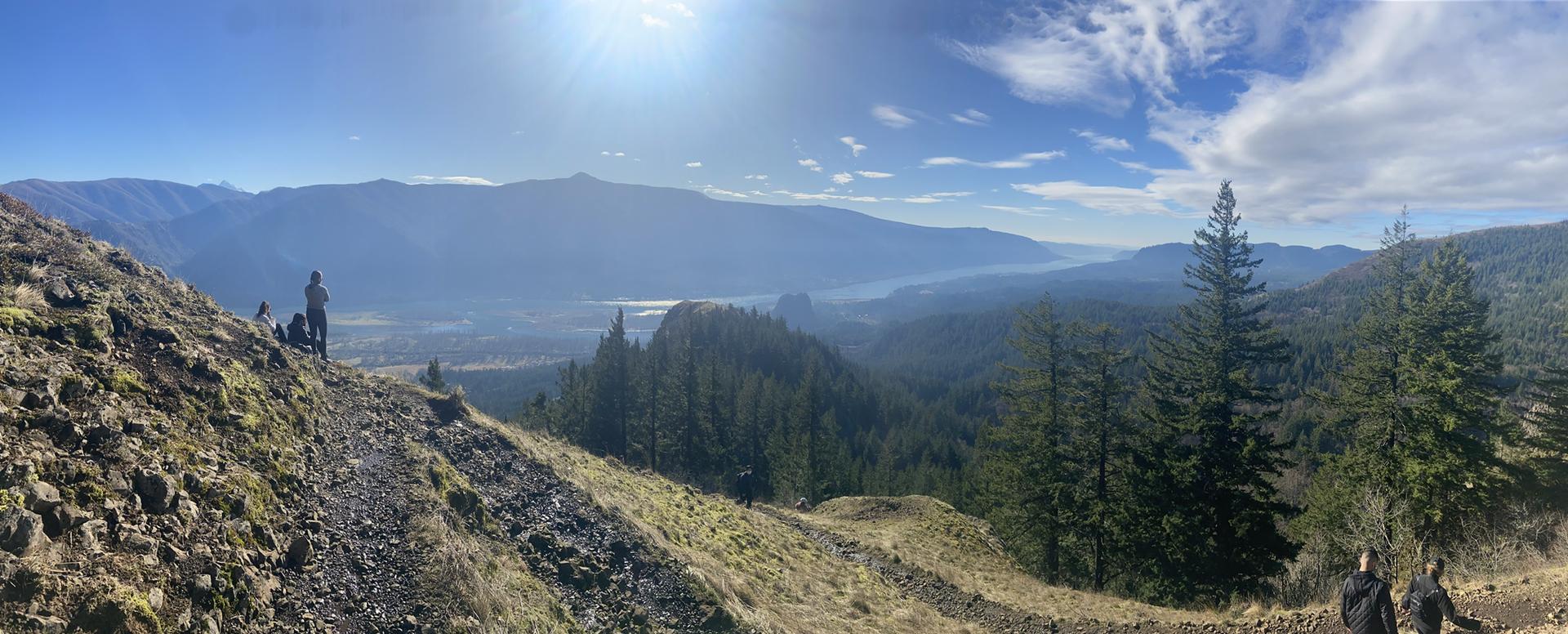 Vast valley, mountains, and forests under bright sun with people on trail.