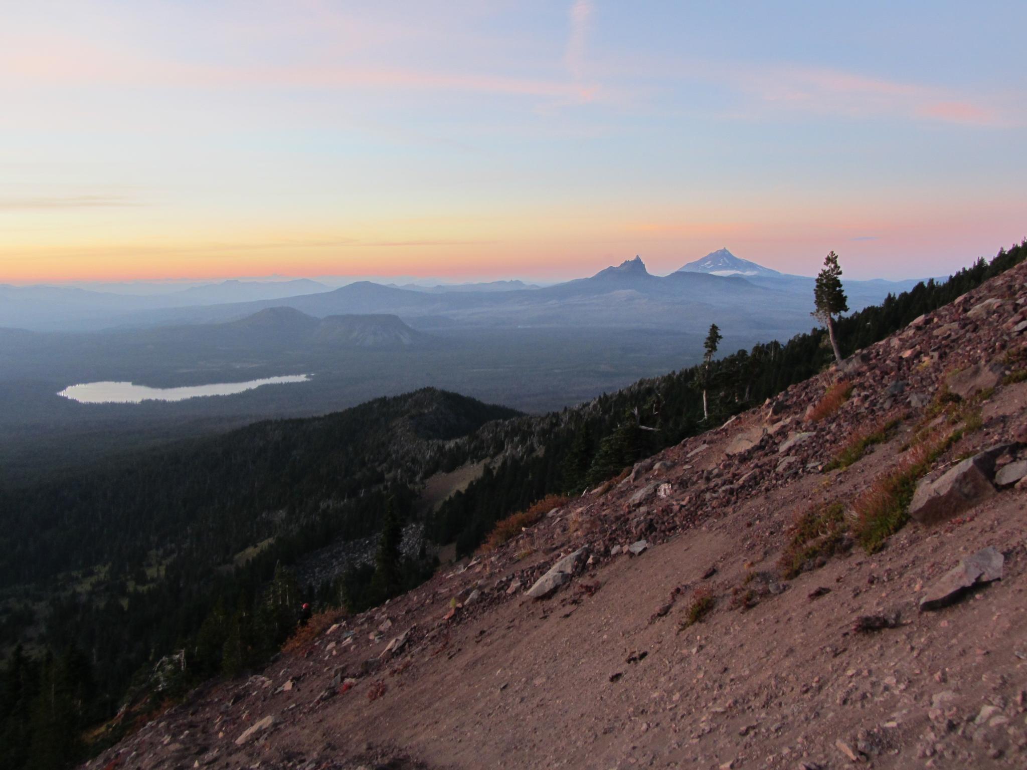 A rocky mountain slope overlooks a distant lake and mountains at sunset.