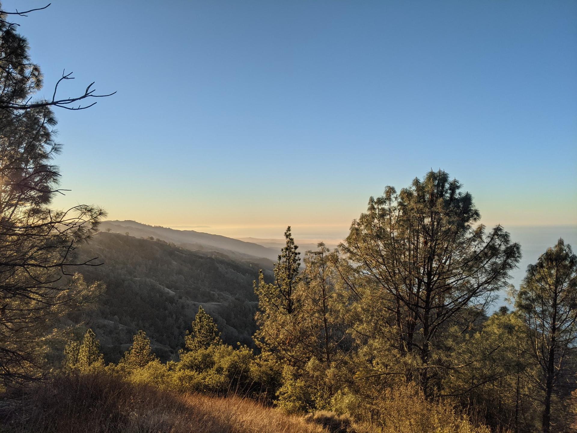 Warm light illuminates forested mountains and a hazy distant horizon at sunset.