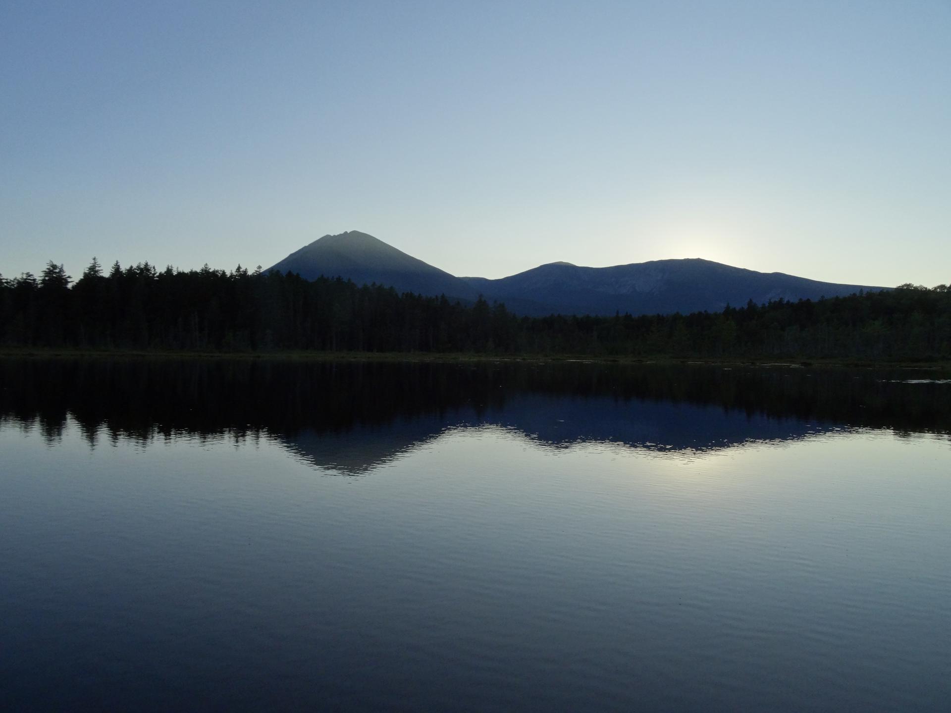 Calm lake reflecting dark mountains and a bright sunrise or sunset.