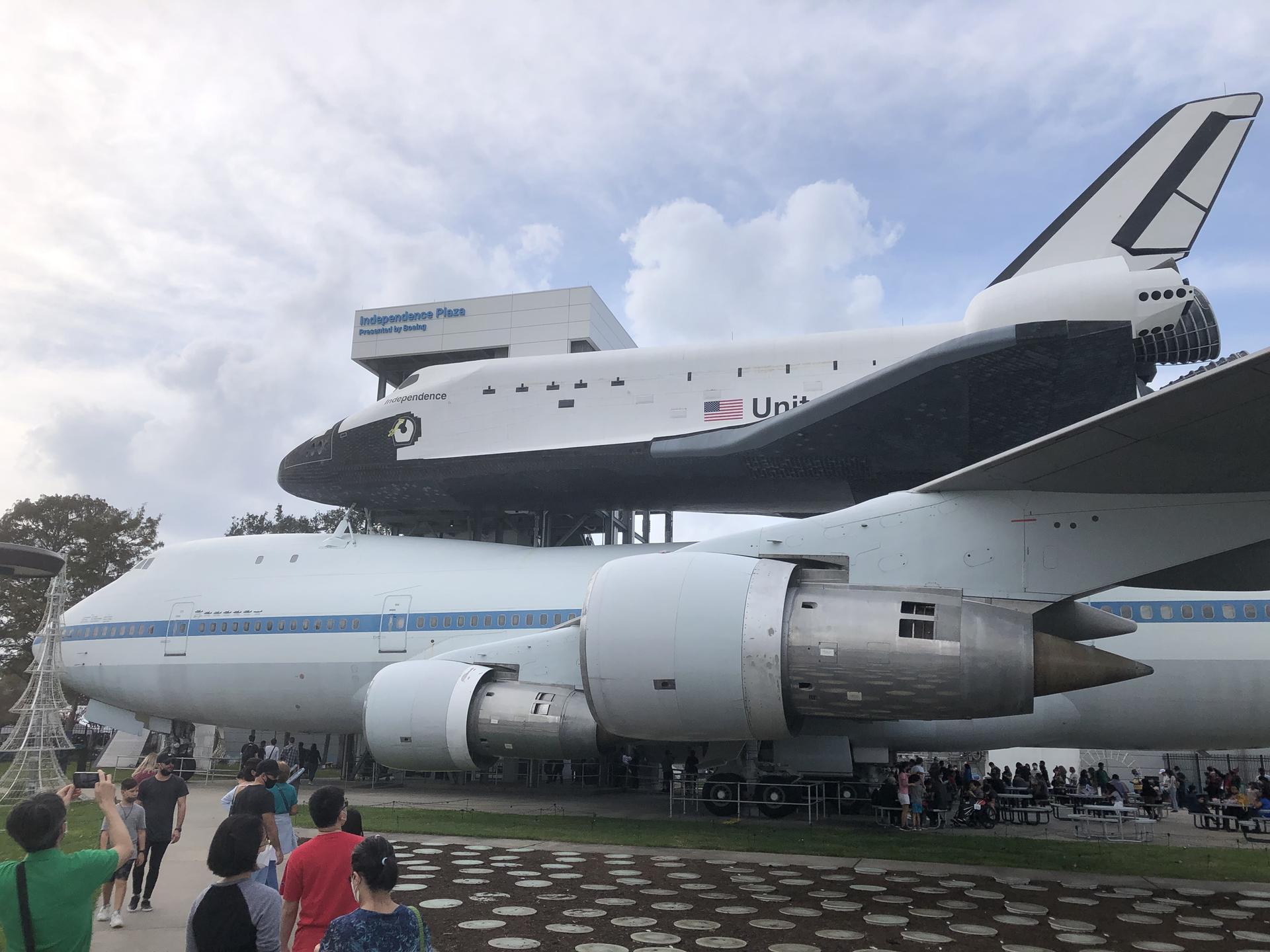 People view a space shuttle mounted on a large white airplane outdoors.