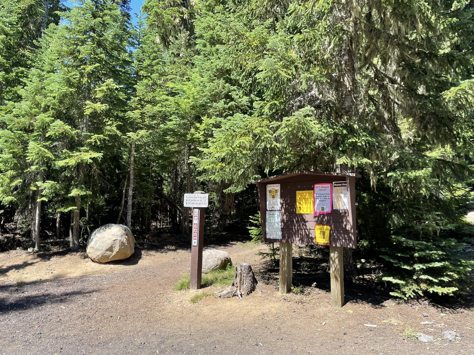 Trailhead with information board, signpost, and large boulder in a dense forest.