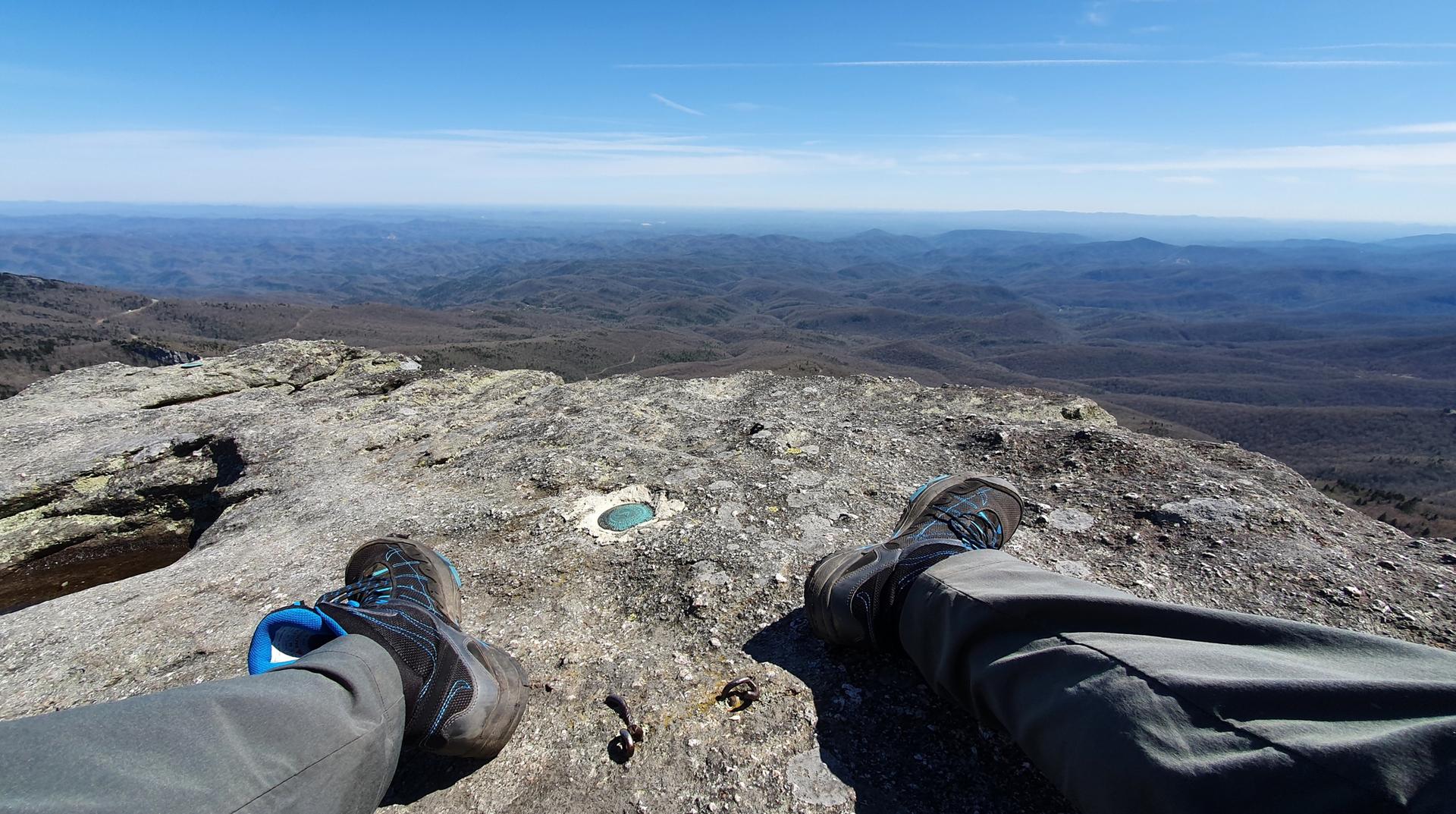 Person's legs and feet resting on a rocky mountain peak with vast landscape.