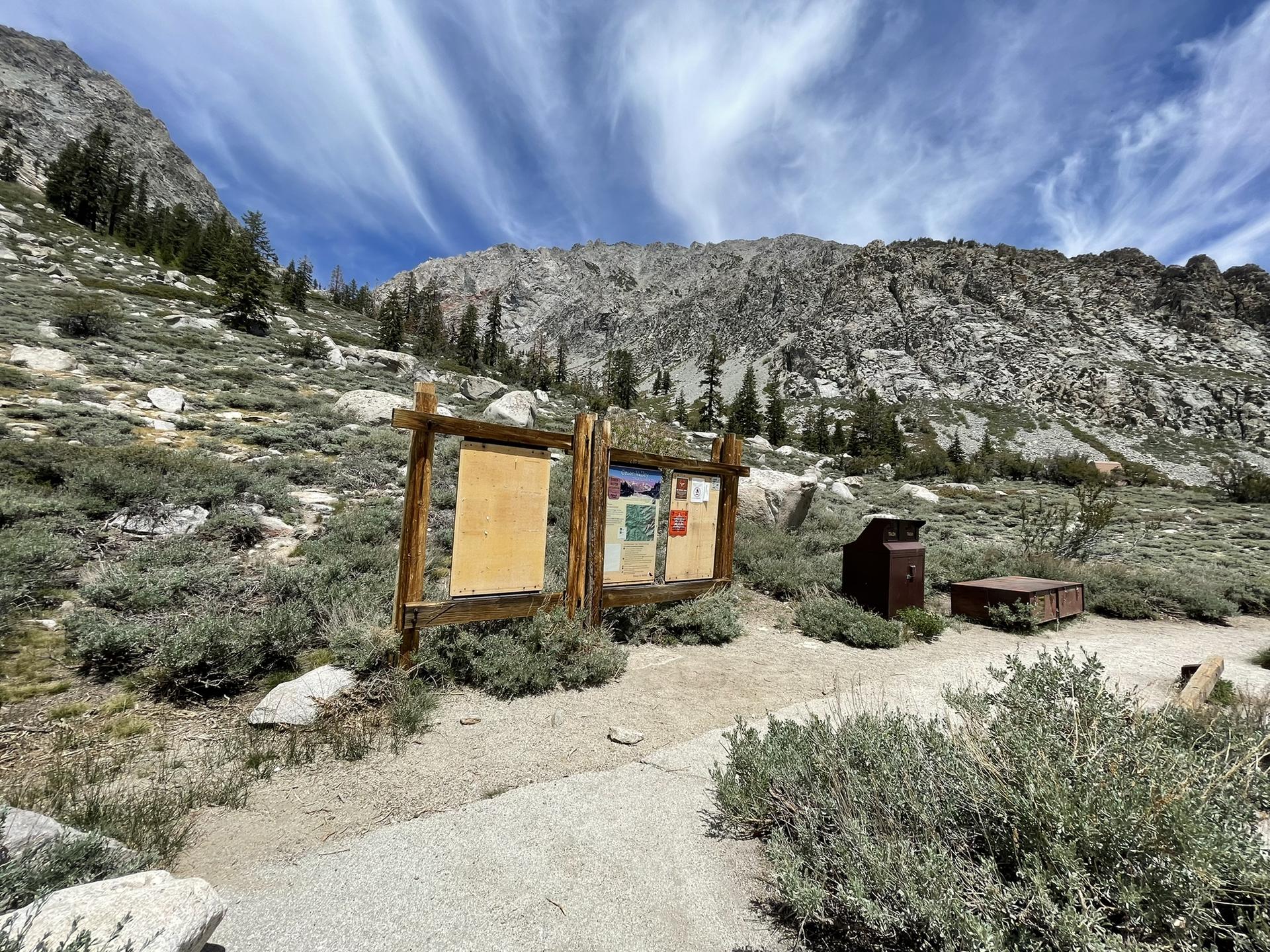 Information boards and trash cans at a mountain trailhead under a cloudy sky.