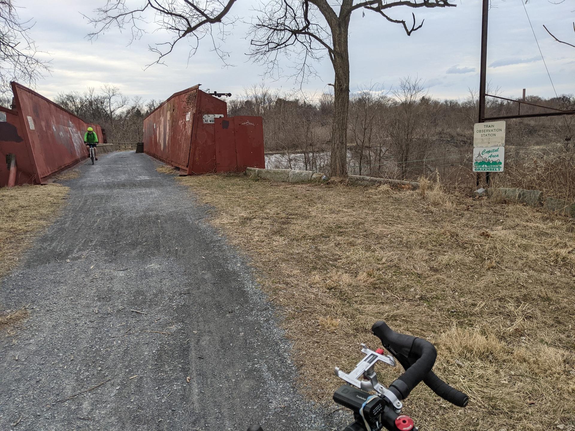 A cyclist on a gravel path next to red industrial structures and bare trees.