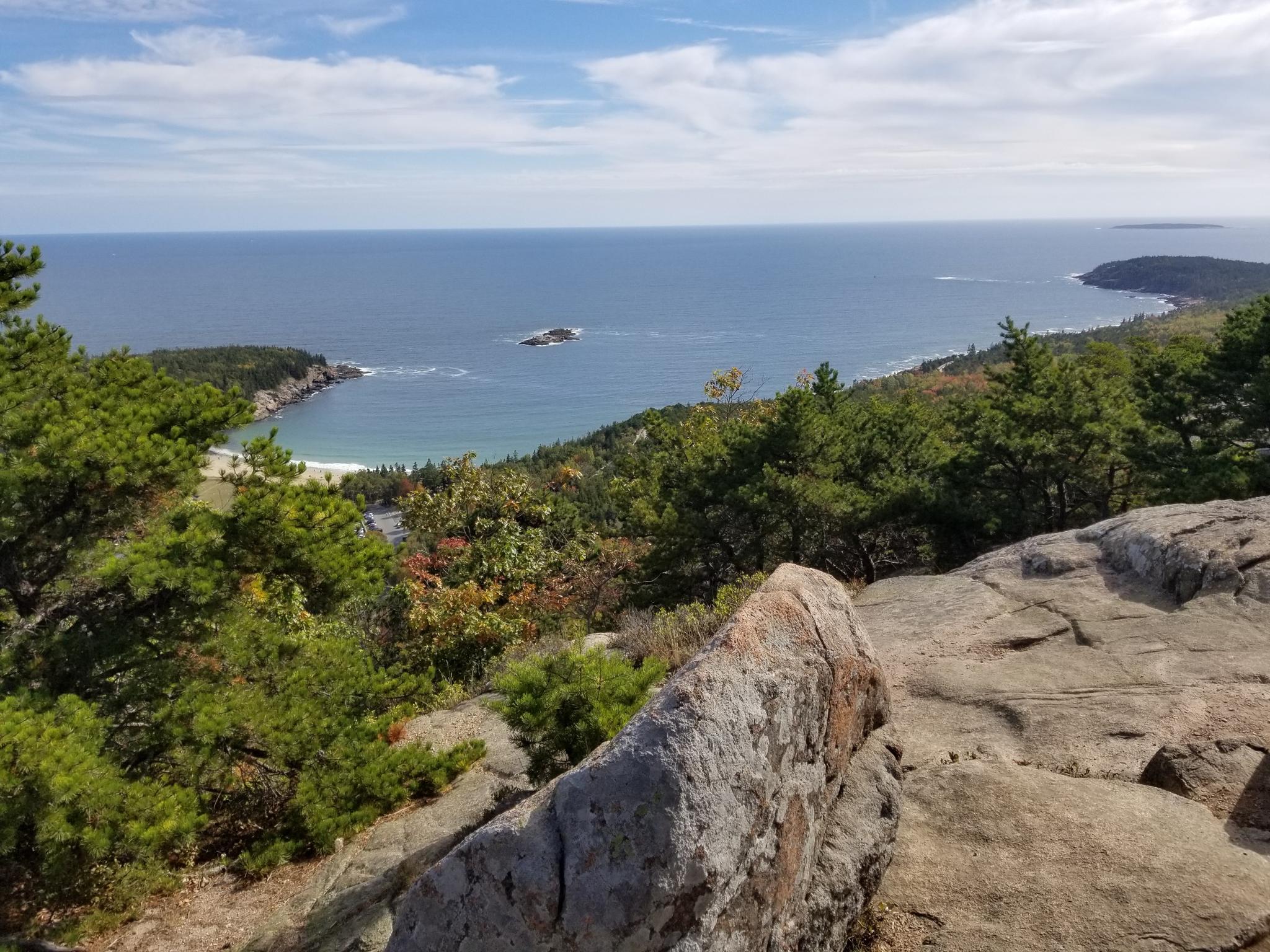 Rocky coastal overlook with green trees, sandy bay, ocean, and cloudy sky.