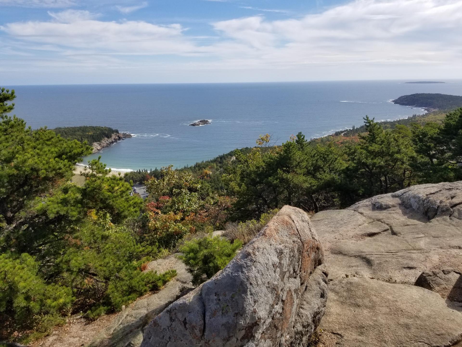 Rocky coastal overlook with green trees, sandy bay, ocean, and cloudy sky.