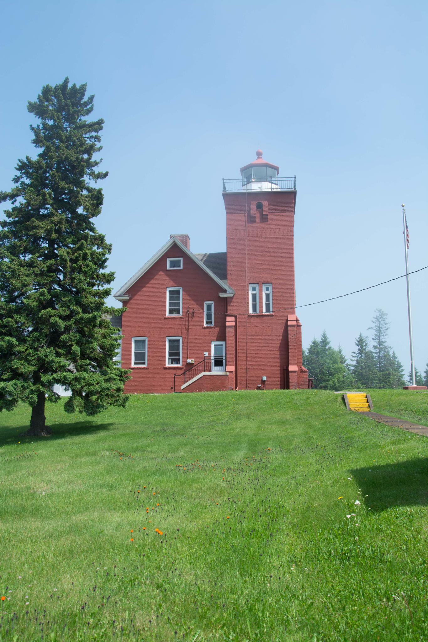 A red brick lighthouse and house on a green grassy hill under a clear sky.