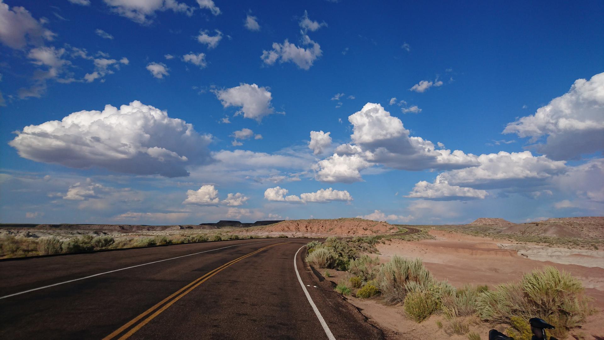 A paved road curves through a desert landscape under a bright, cloudy sky.
