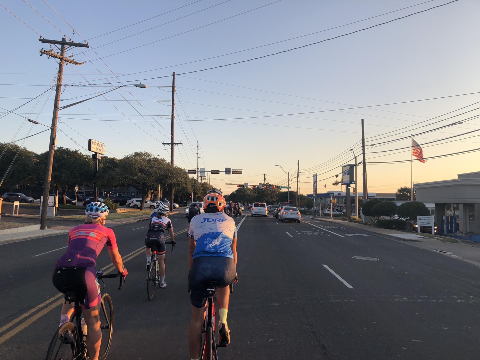 Three cyclists ride on a sunny urban road with power lines overhead.